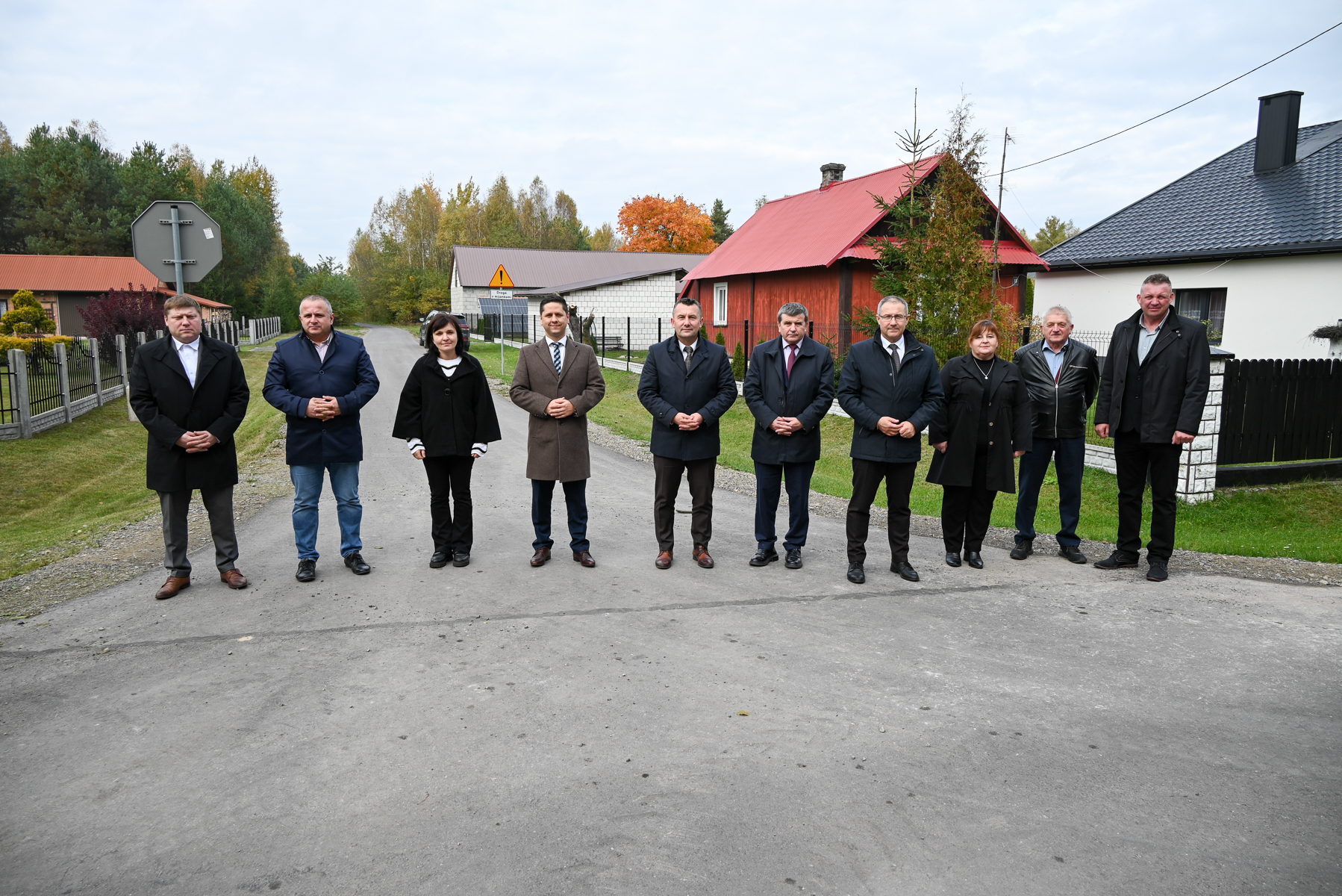 Dziesięć osób w płaszczach i kurtkach stoi na drodze między domami z ogrodzeniami, w tle drzewa o jesiennych kolorach.