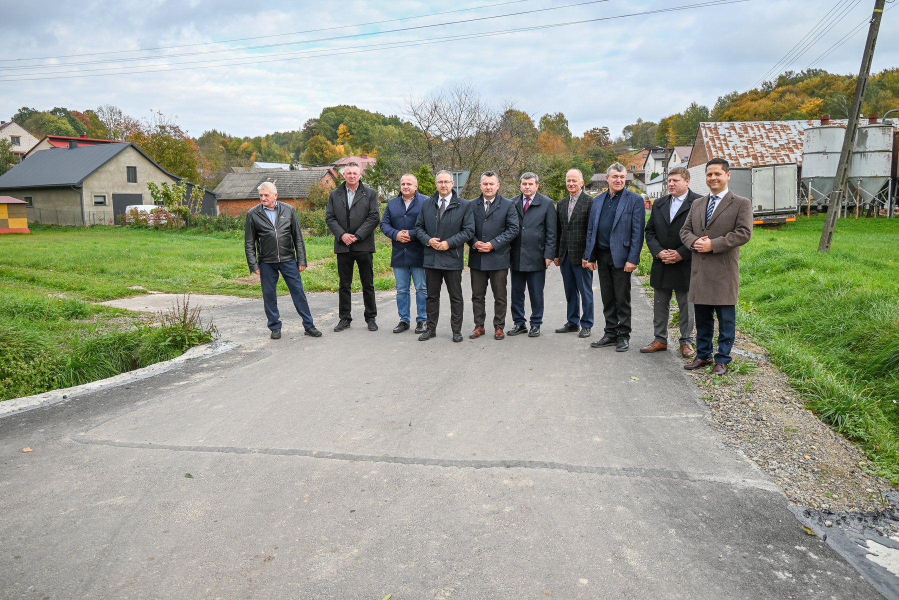 Dziesięciu mężczyzn w jesiennych płaszczach stoi na asfaltowej drodze, wokół zielona trawa, budynki wiejskie, drzewa z kolorowymi liśćmi i pochmurne niebo.