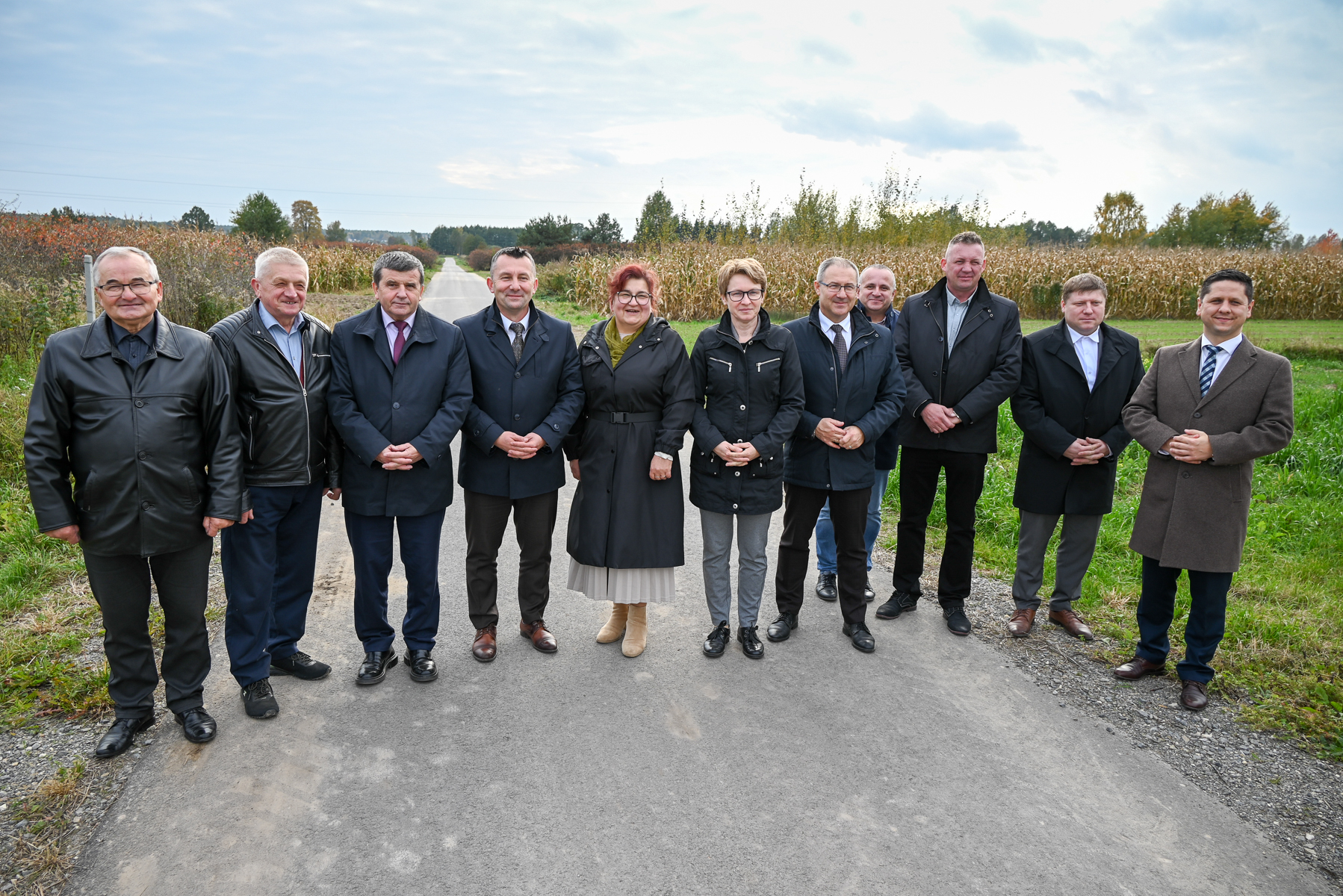 Grupa jedenastu osób w jesiennych płaszczach stoi na asfaltowej drodze na tle pól kukurydzy i drzew pod pochmurnym niebem.