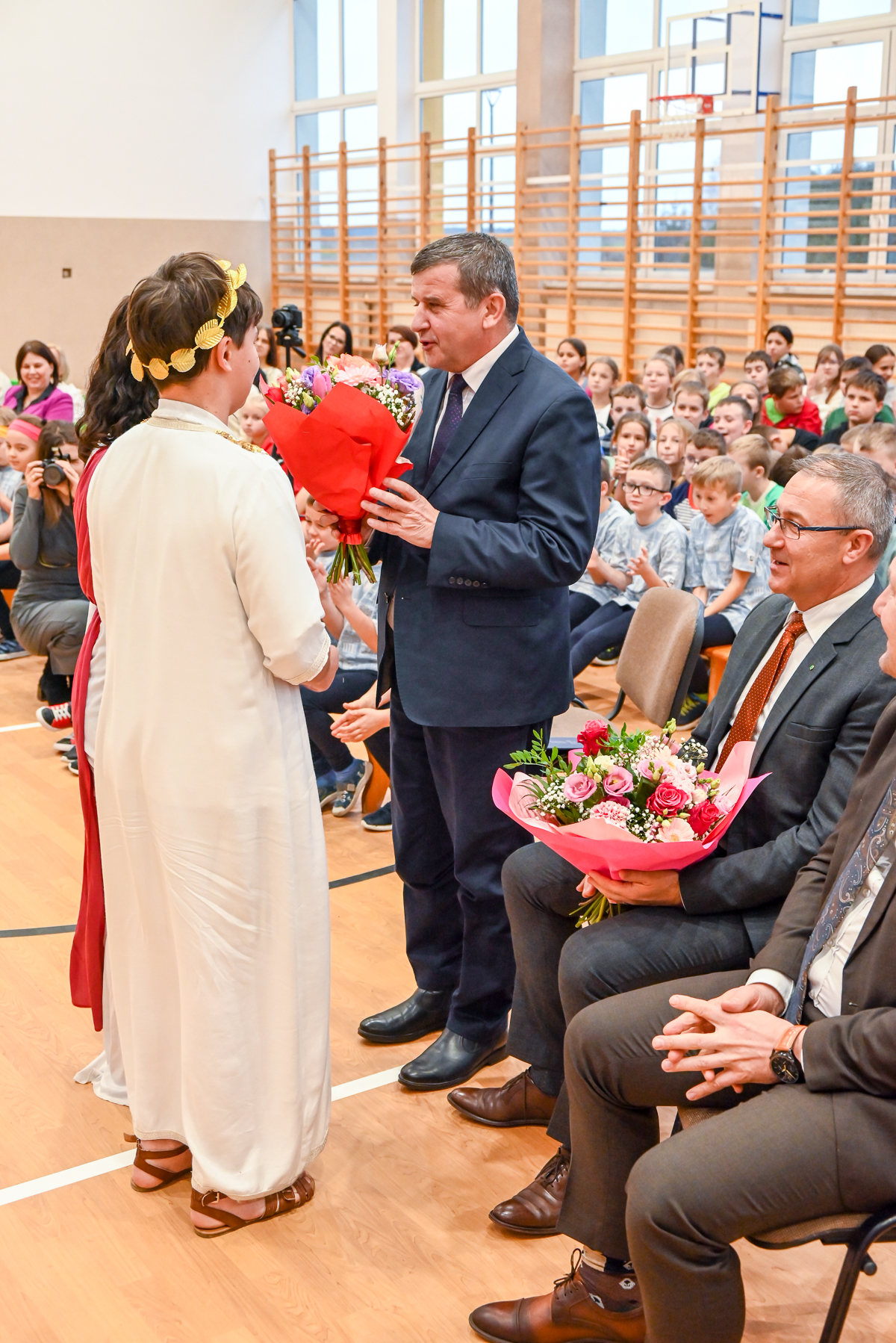 Mężczyzna w granatowym garniturze wręcza bukiet kwiatów osobie w długiej białej szacie i złotym wieńcu, w tle dzieci i dorośli siedzący na sali gimnastycznej.