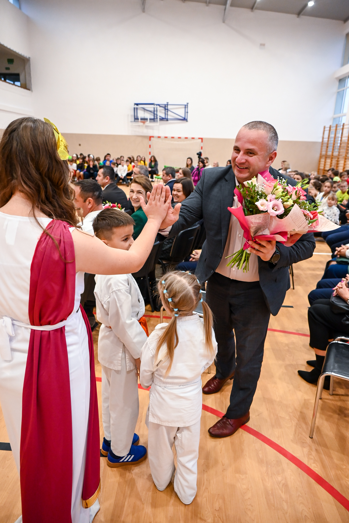 Mężczyzna w garniturze z bukietem kwiatów daje piątkę kobiecie w białej sukience z czerwonym szalem, obok dzieci w białych strojach sportowych, w tle publiczność na sali gimnastycznej.