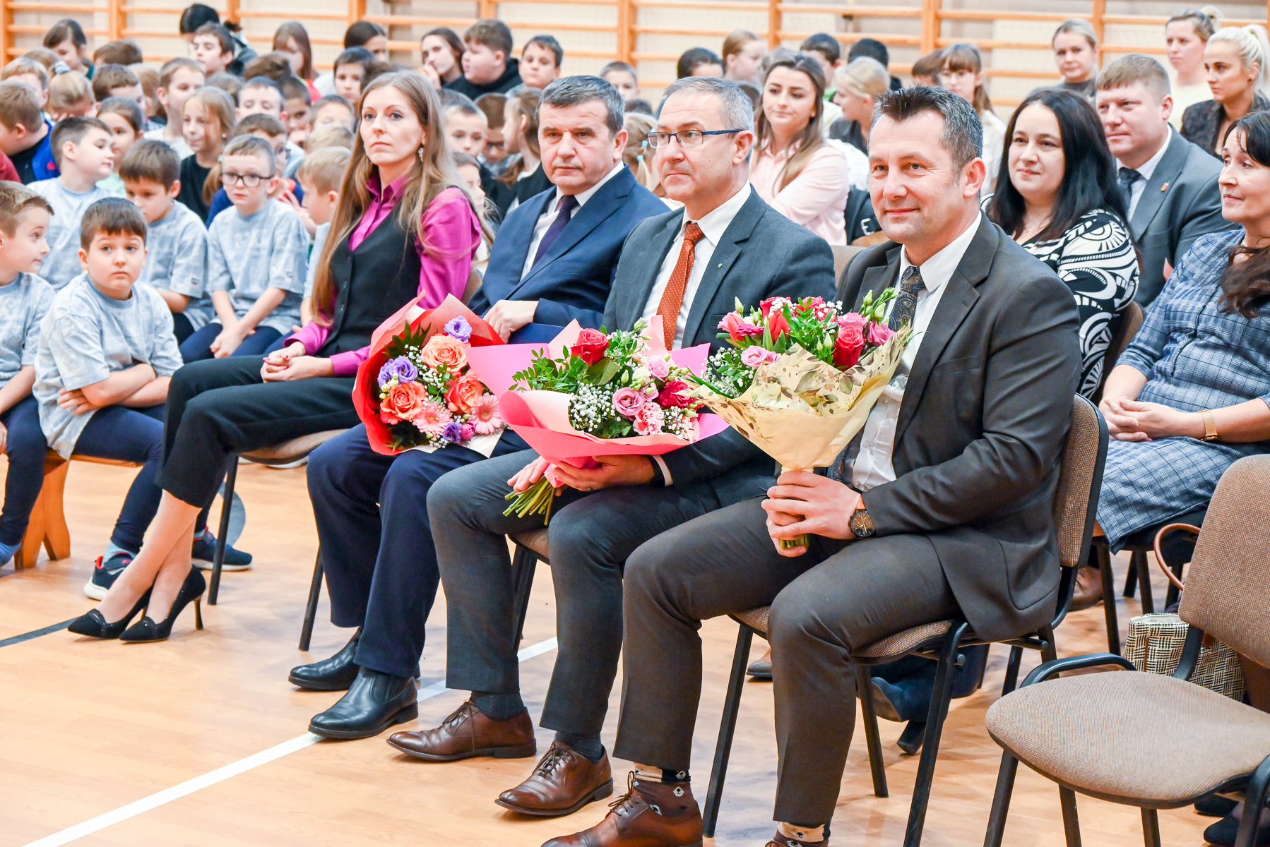 Grupa osób siedzących na krzesłach w sali gimnastycznej, trzymających bukiety kwiatów, w tle dzieci w szarych koszulkach i dorośli.