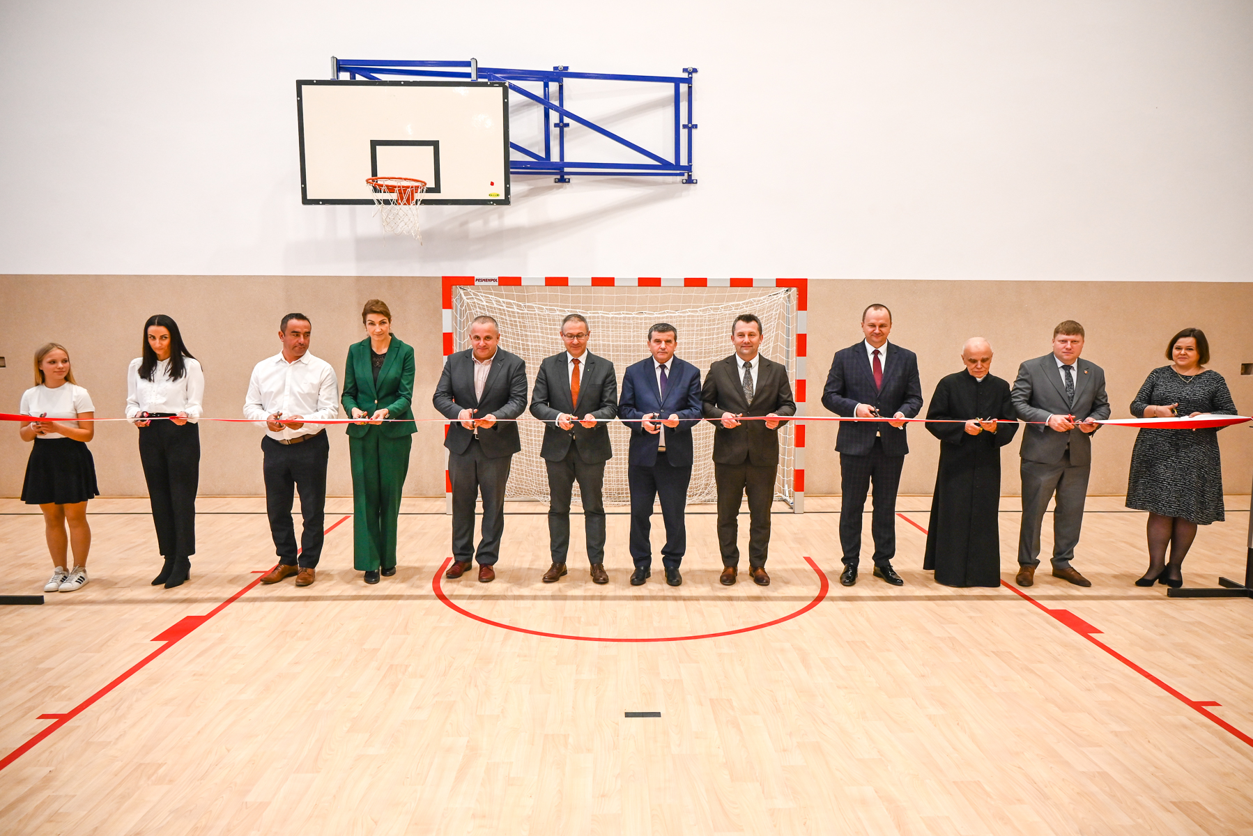 Dwunastu ludzi stoi w hali sportowej na drewnianej podłodze przed bramką do piłki ręcznej, trzymając czerwoną wstęgę do przecięcia, nad nimi kosz do koszykówki.