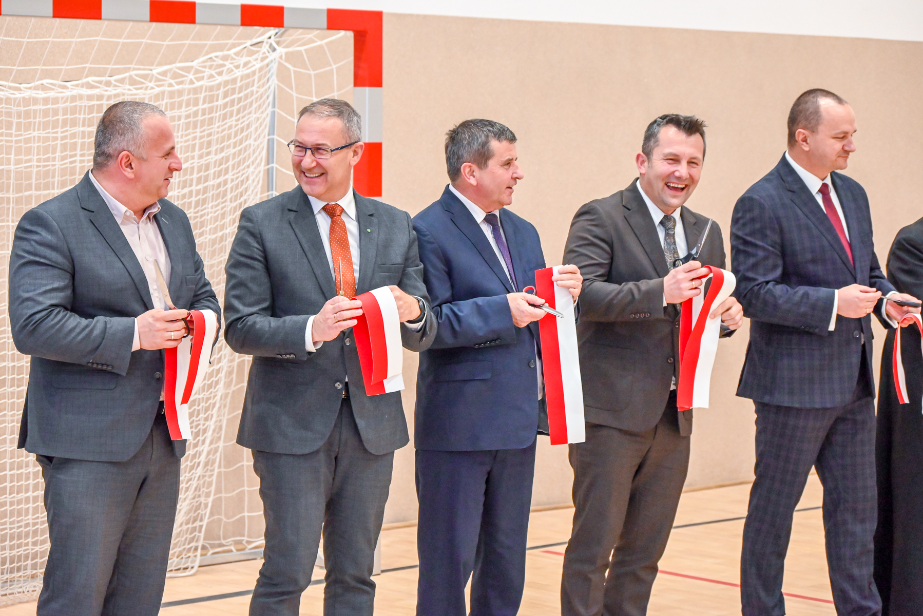 Pięciu mężczyzn w garniturach trzyma nożyczki i biało-czerwone wstęgi na tle bramki sportowej i jasnej ściany.