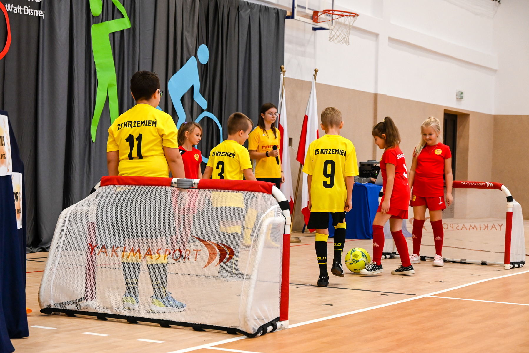 Dzieci w żółtych i czerwonych strojach sportowych stoją na sali gimnastycznej przy dwóch małych bramkach do piłki nożnej, na parkiecie żółta piłka.