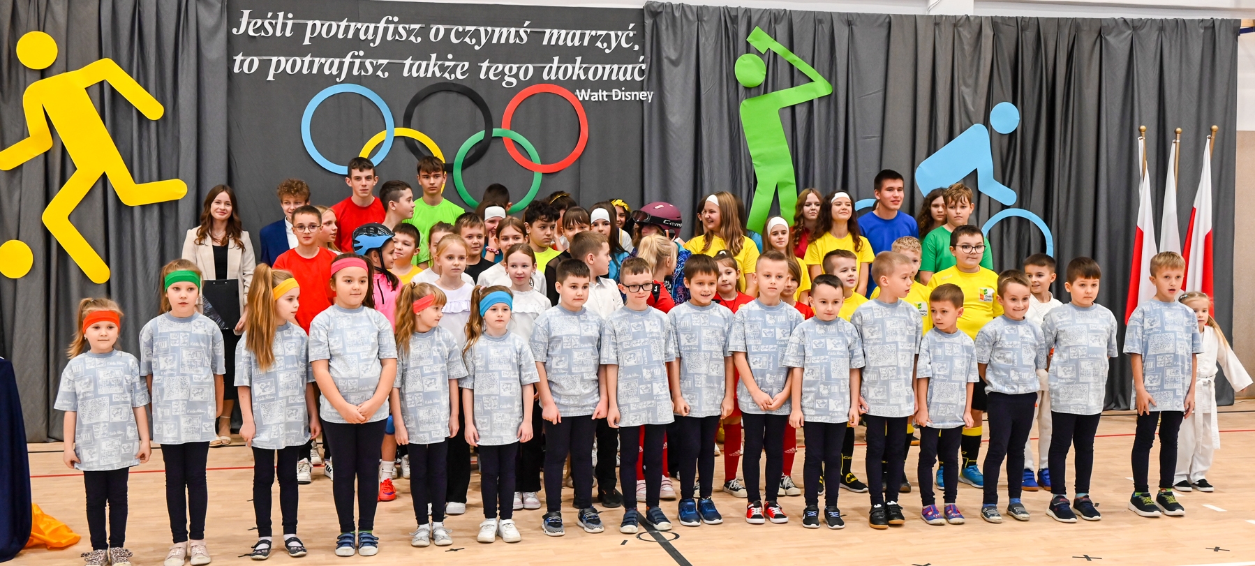 Grupa dzieci i nastolatków w kolorowych strojach sportowych stoi na tle szarej kurtyny z olimpijskimi pierścieniami i dużymi kolorowymi sylwetkami sportowców.