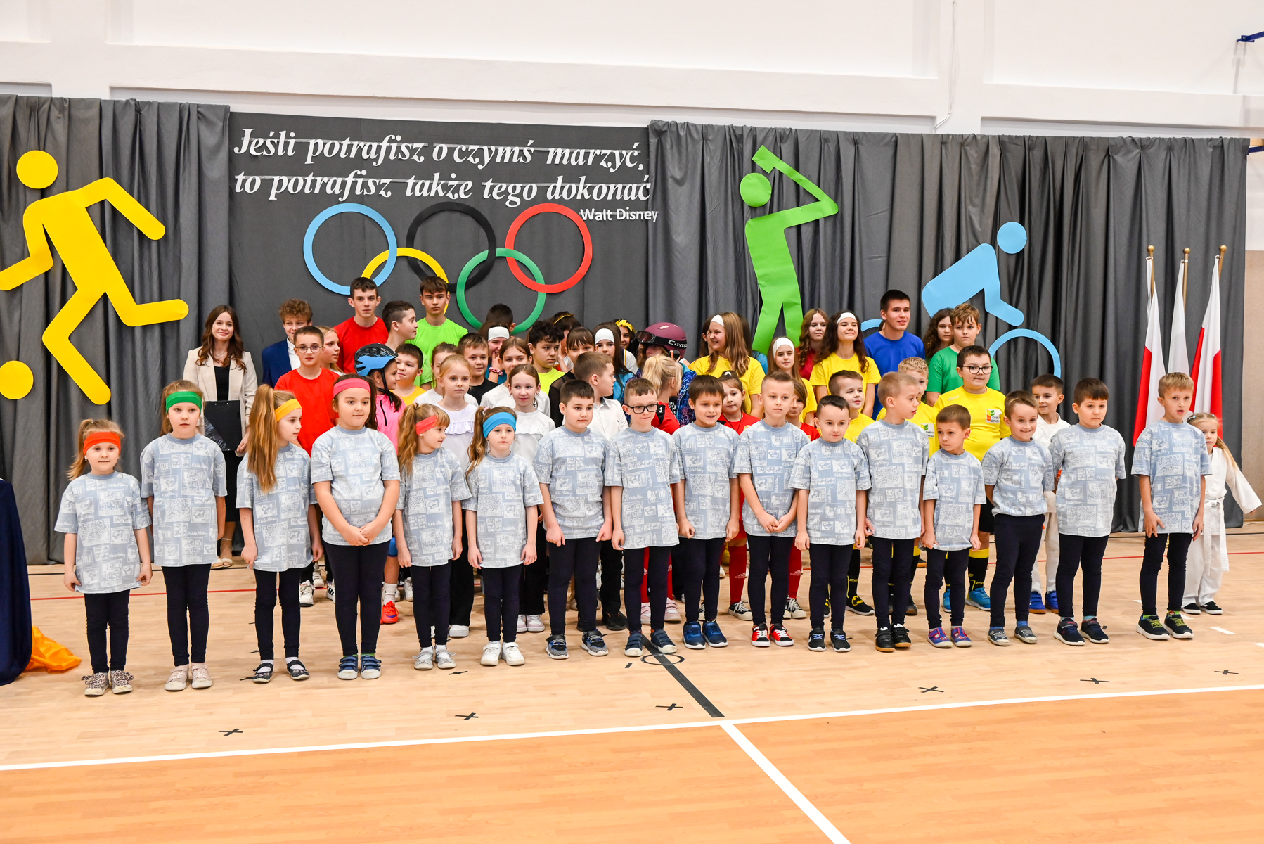 Grupa dzieci i młodzieży w kolorowych strojach sportowych stoi na sali gimnastycznej przed szarą kurtyną z kolorowymi symbolami sportów i cytatem Walt Disney, po prawej trzy flagi Polski.
