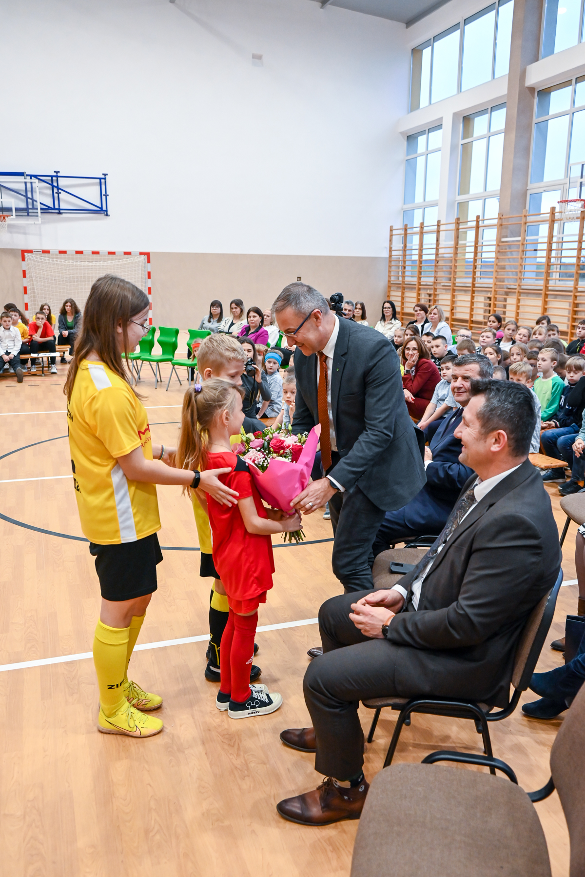 Mężczyzna w garniturze odbiera bukiet róż od dzieci w sportowych strojach na sali gimnastycznej, w tle siedzą widzowie na trybunach i krzesłach.
