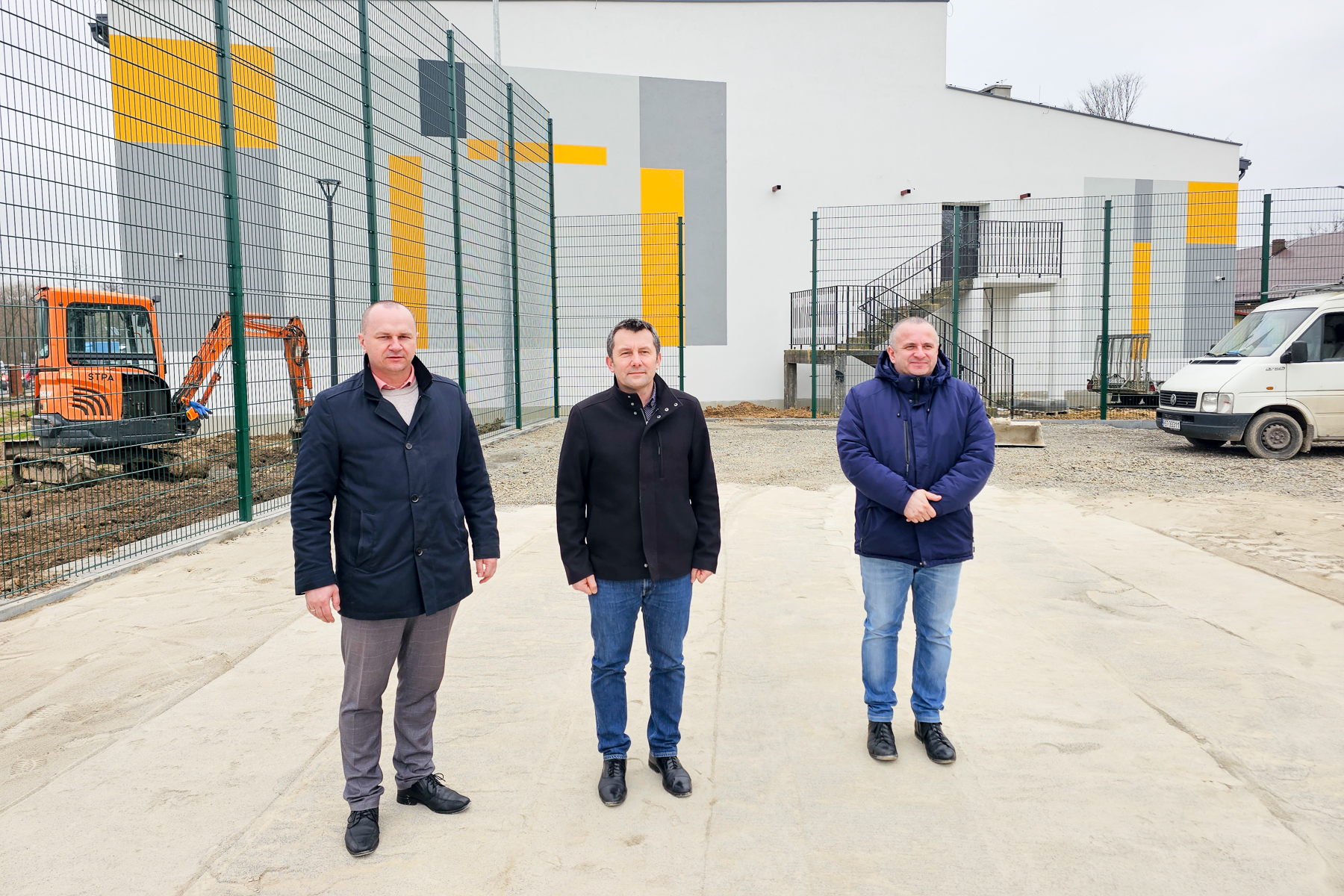 Trzech mężczyzn w kurtkach stoi na jasnym placu przed ogrodzeniem i budynkiem z szaro-żółtymi panelami, obok biały van i pomarańczowa koparka.