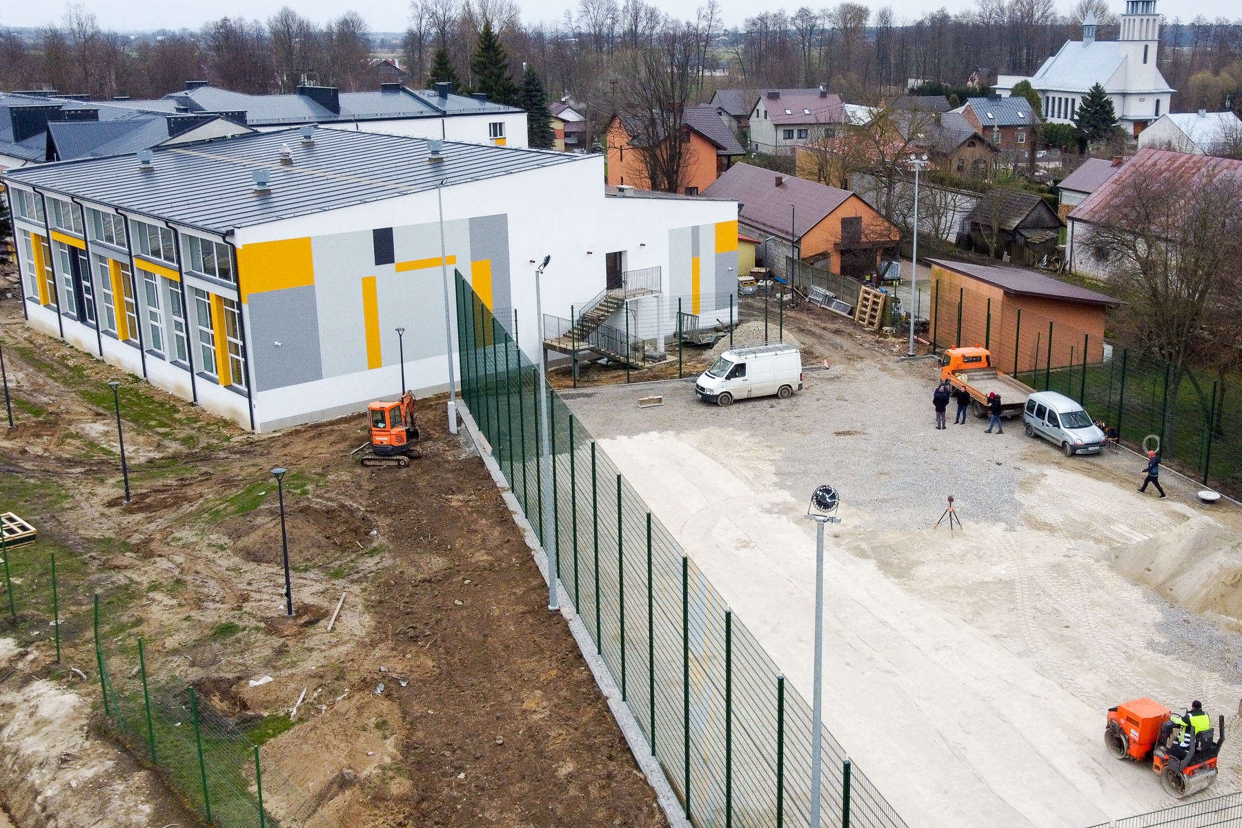 Budynek z żółto-szarymi pasami, plac budowy z maszyną budowlaną, ogrodzenie, samochody i kilka osób na terenie, w tle domy i kościół.