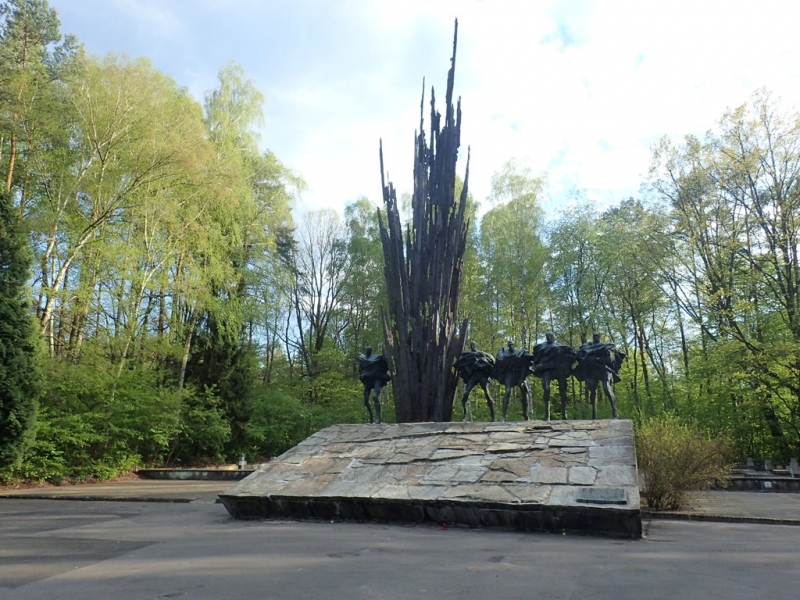 Pomnik z postaciami jeźdźców na koniach z czarnym, wysokim, ostrym elementem, na kamiennym cokole, w otoczeniu zielonych drzew i niebieskiego nieba.