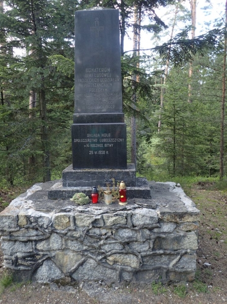 Czarny kamienny pomnik z napisem na kamiennej podstawie, otoczony drzewami iglastymi, z trzema zniczami i kwiatami u podstawy.