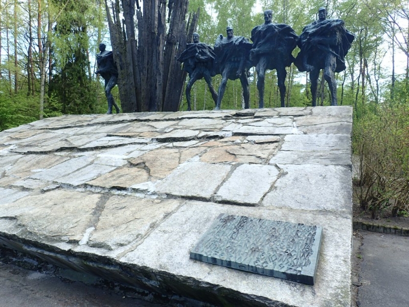 Pomnik z sześcioma ciemnymi, stylizowanymi postaciami na dużej, kamiennej podstawie, otoczony zielonymi drzewami i krzewami.