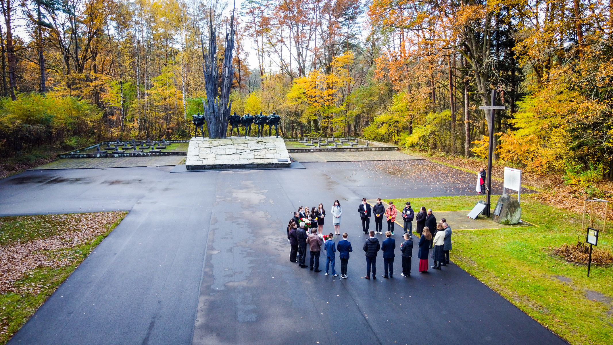 Grupa ludzi stojąca w kole na ciemnym asfalcie obok dużego krzyża i pomnika z czarnymi rzeźbami na tle jesiennego lasu.