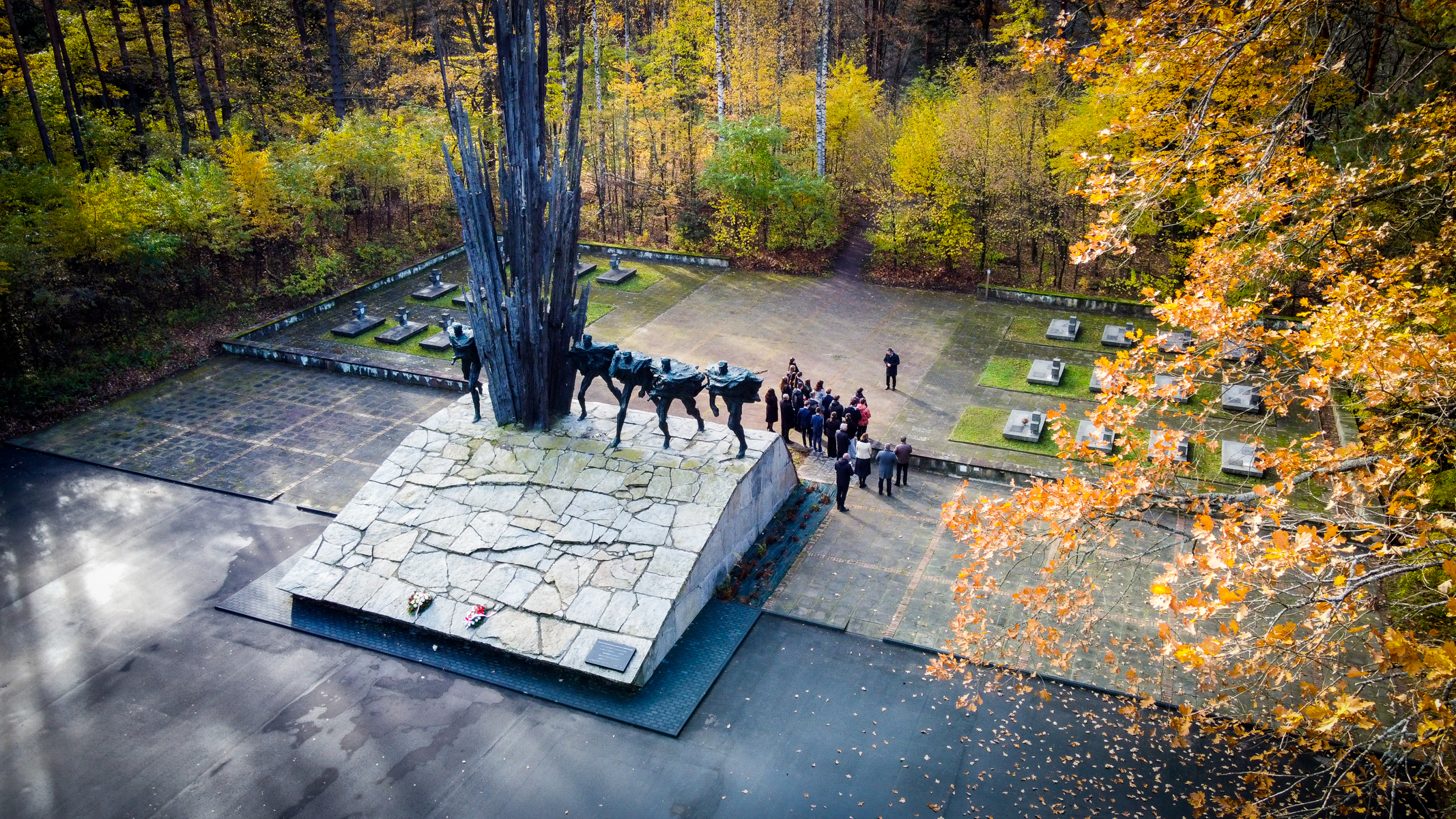 Pomnik z postaciami na kamiennej podstawie na tle jesiennego lasu, w pobliżu grupa ludzi i nagrobki w zielonych kwadratach, złote liście na drzewach.