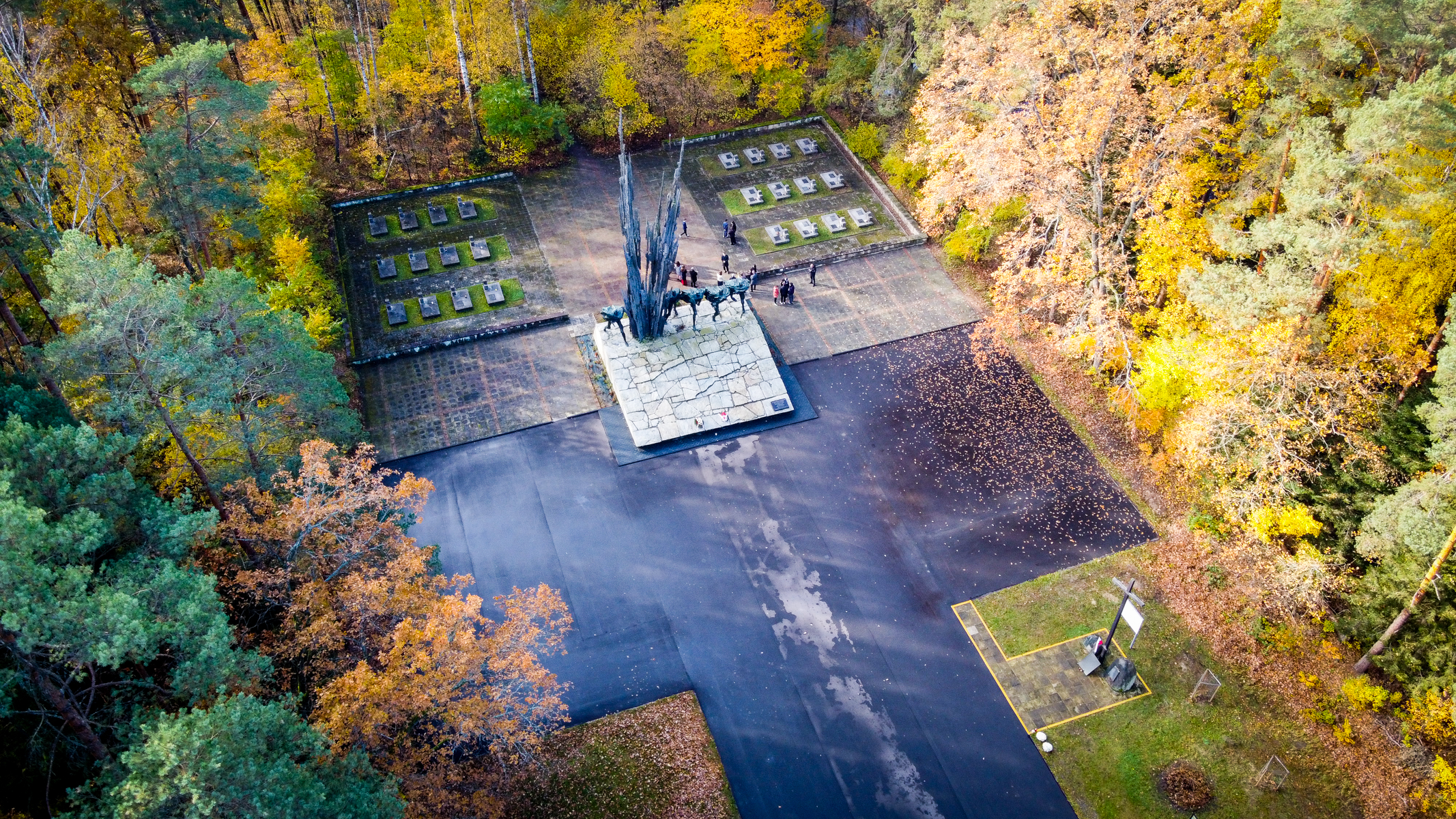 Czarny pomnik z postaciami i wysokimi kolcami na kamiennej podstawie, otoczony alejkami i kolorowymi jesiennymi drzewami. Grupa ludzi przy pomniku.