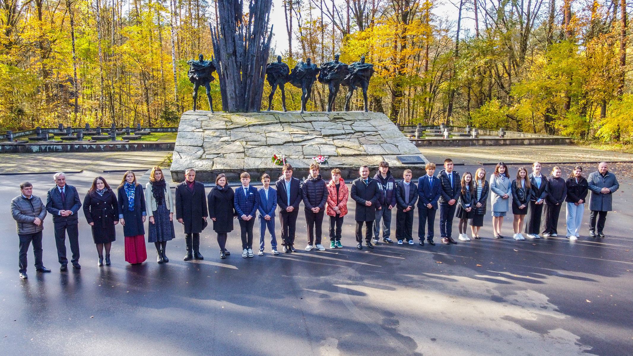 Grupa ludzi stoi na tle pomnika z postaciami na kamiennej podstawie, otoczony jesiennym lasem z żółtymi i zielonymi liśćmi.