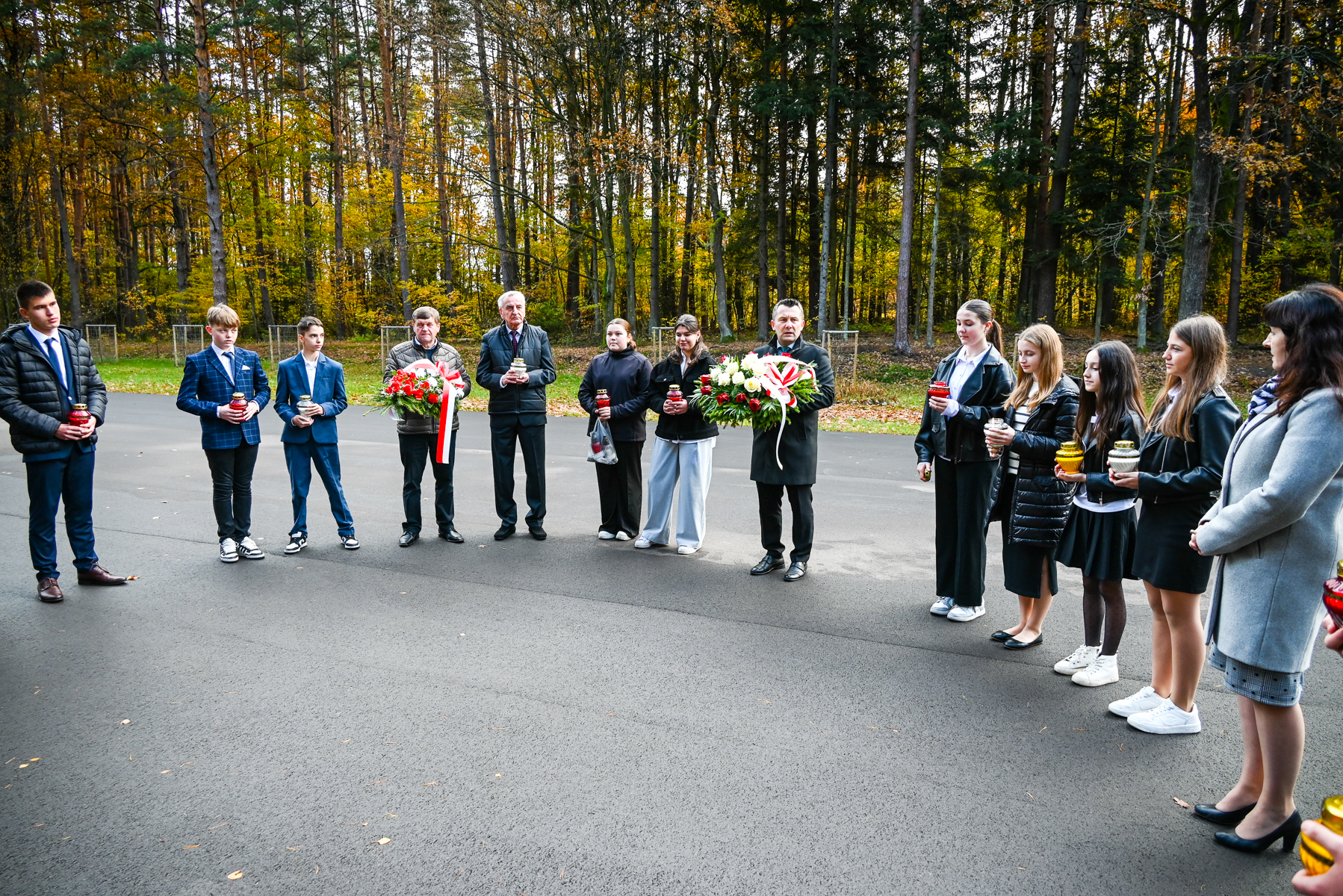 Grupa ludzi stoi na asfaltowej drodze w półokręgu, trzymając znicze i dwa wieńce z czerwono-białymi wstążkami, w tle las z drzewami o jesiennych kolorach liści.