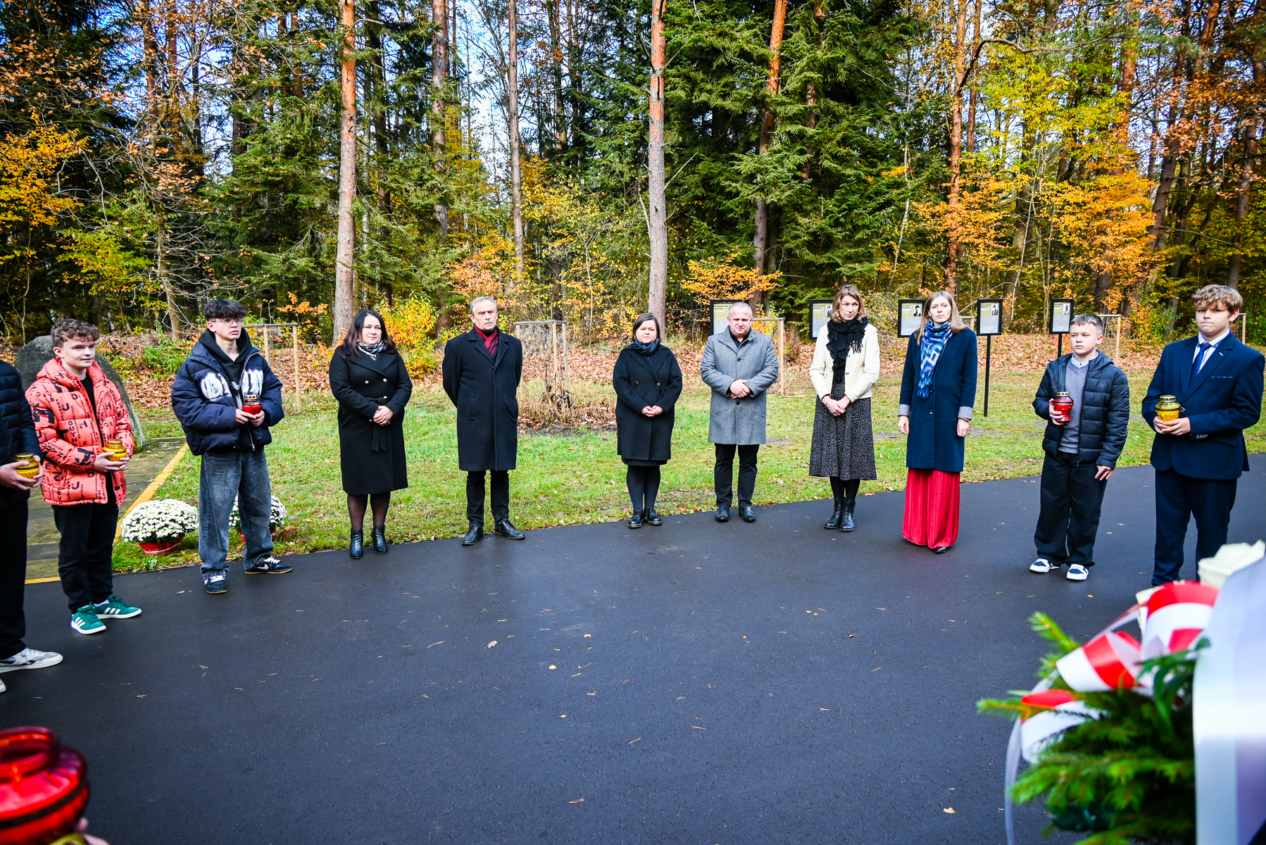 Grupa osób stojących na czarnej nawierzchni na tle jesiennego lasu, kilka osób trzyma czerwone i żółte znicze. Po prawej zielone gałązki z biało-czerwoną wstążką.