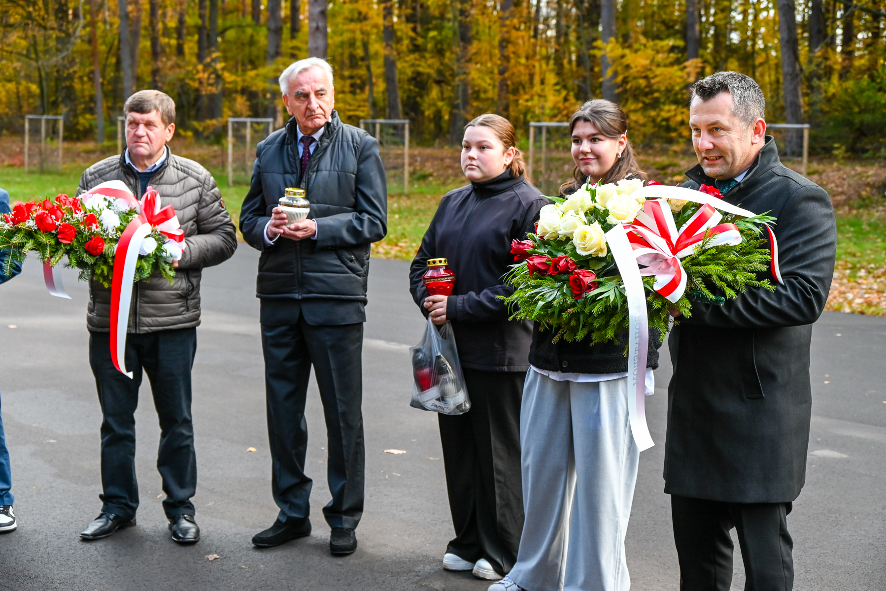 Grupa pięciu osób stoi na asfaltowej ścieżce, dwie trzymają wieńce z czerwono-białymi wstążkami, jedna trzyma plastikową torbę z lampionami, dwie trzymają lampiony, jesienne drzewa w tle.