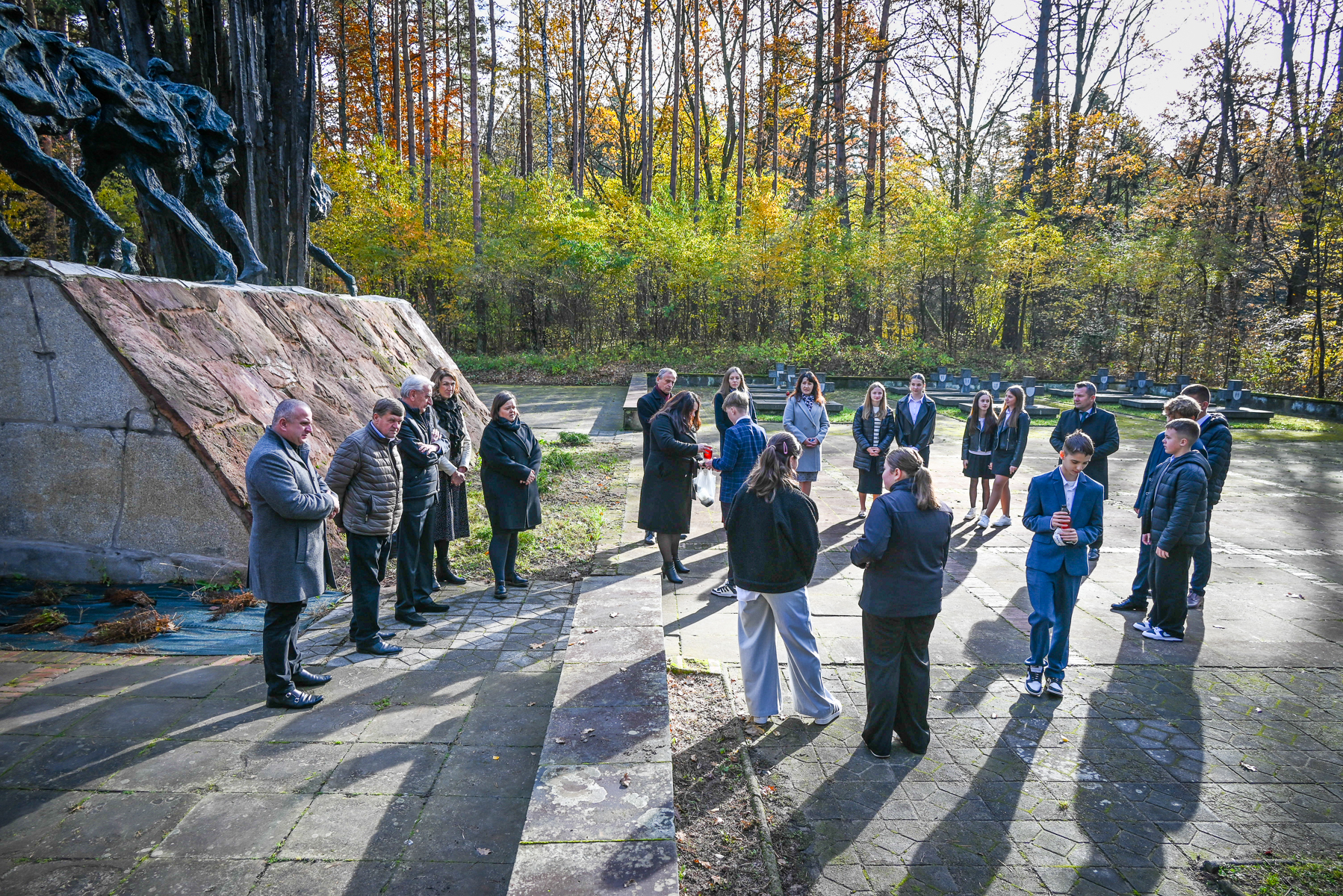 Grupa ludzi stojących na kamiennym placu przy pomniku z ciemnych figur, w tle jesienne drzewa z żółto-zielonymi liśćmi.