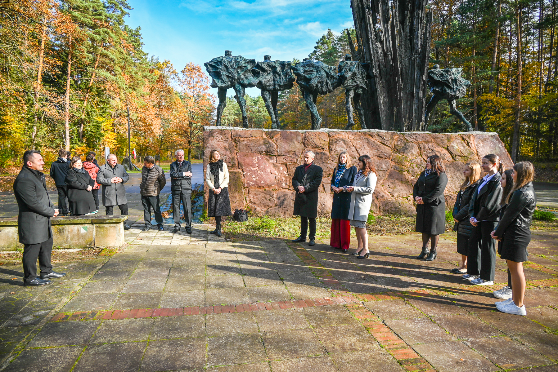 Grupa ludzi stojąca na placu z kamiennym pomnikiem postaci bez głów na tle jesiennych drzew i niebieskiego nieba.