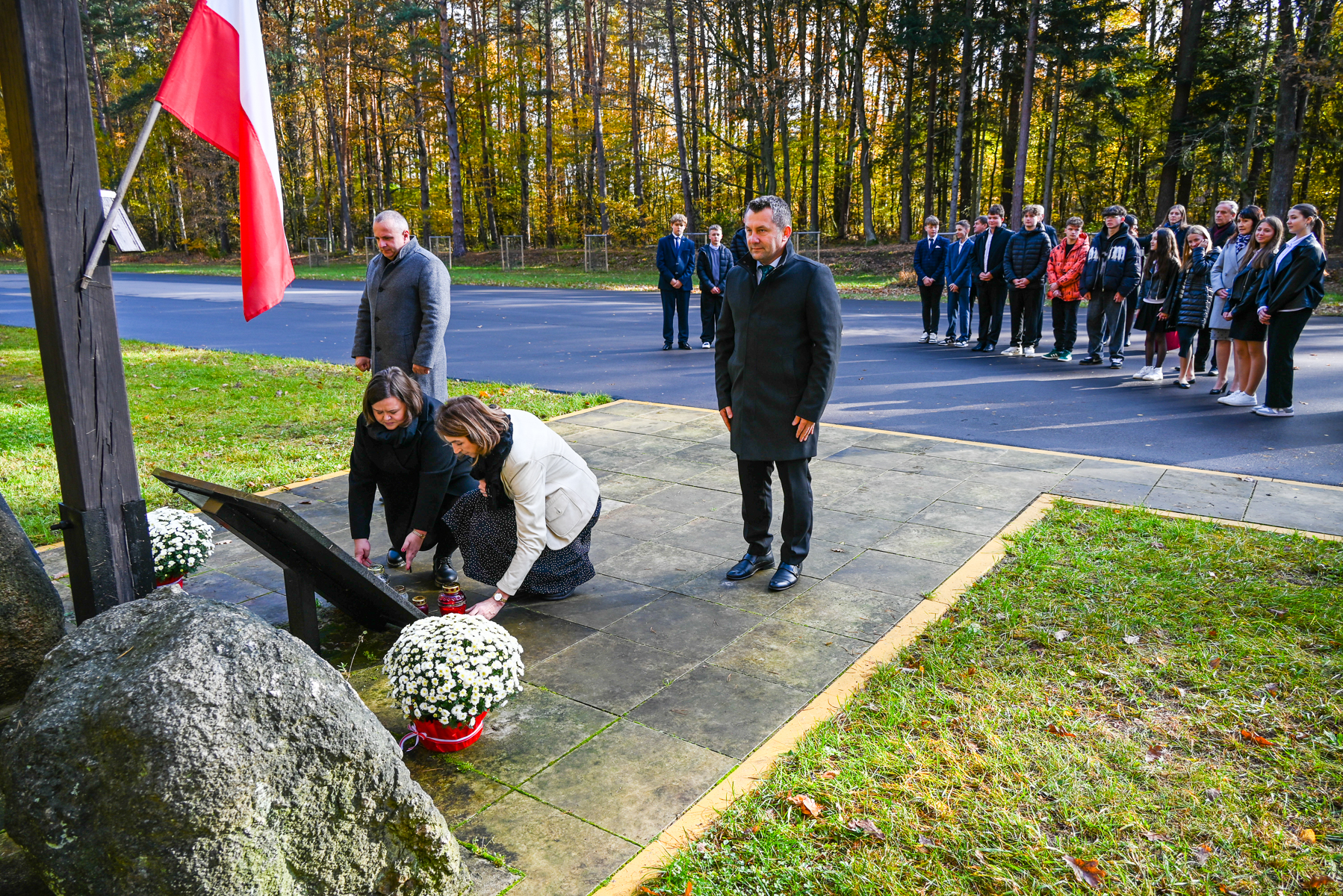 Grupa ludzi na zewnątrz, dwie kobiety kładą znicze na pomniku z tablicą, mężczyzna w płaszczu stoi obok, w tle Polskie flagi i las jesiennych drzew.