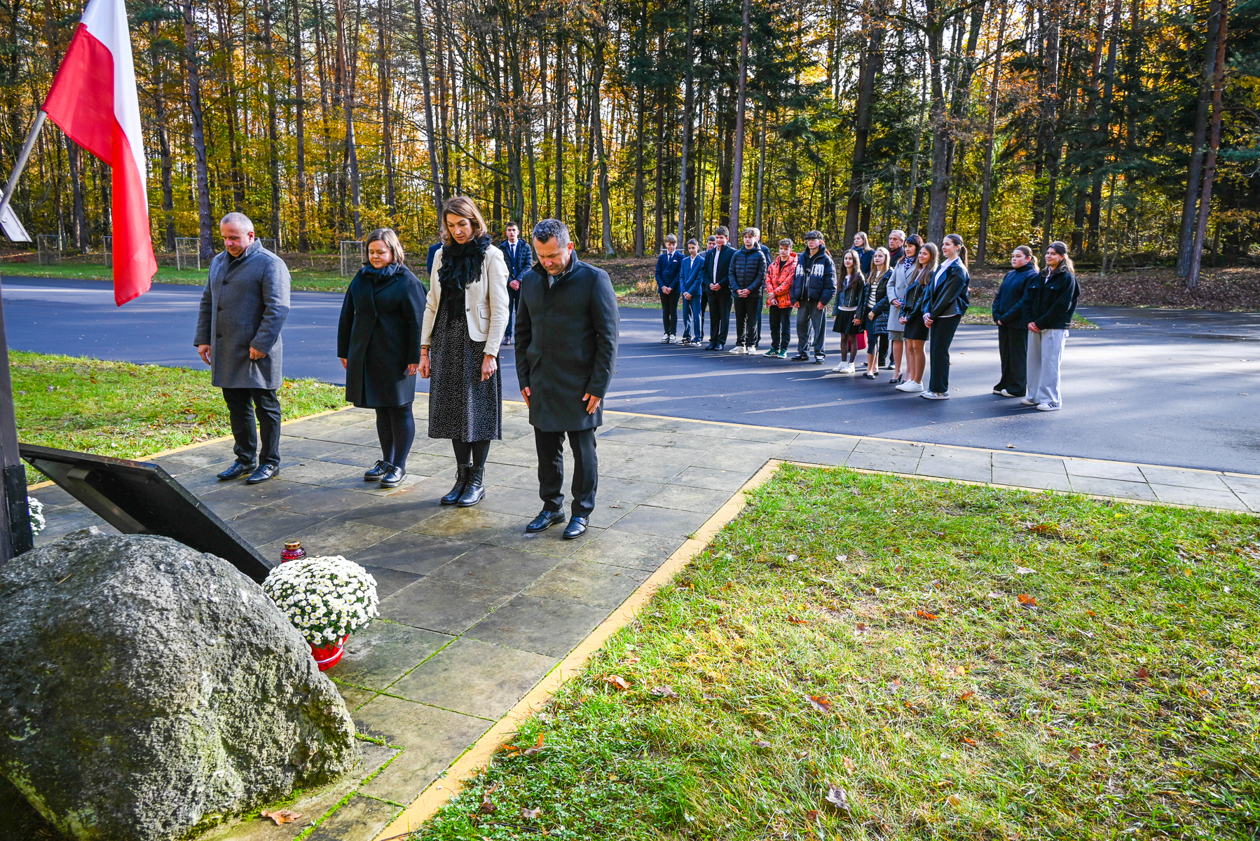 Grupa ludzi w jesiennym parku, cztery osoby z przodu stoją ze spuszczonymi głowami przy pomniku z białymi kwiatami i zniczem, za nimi stoi kilkanaście osób w rzędzie.