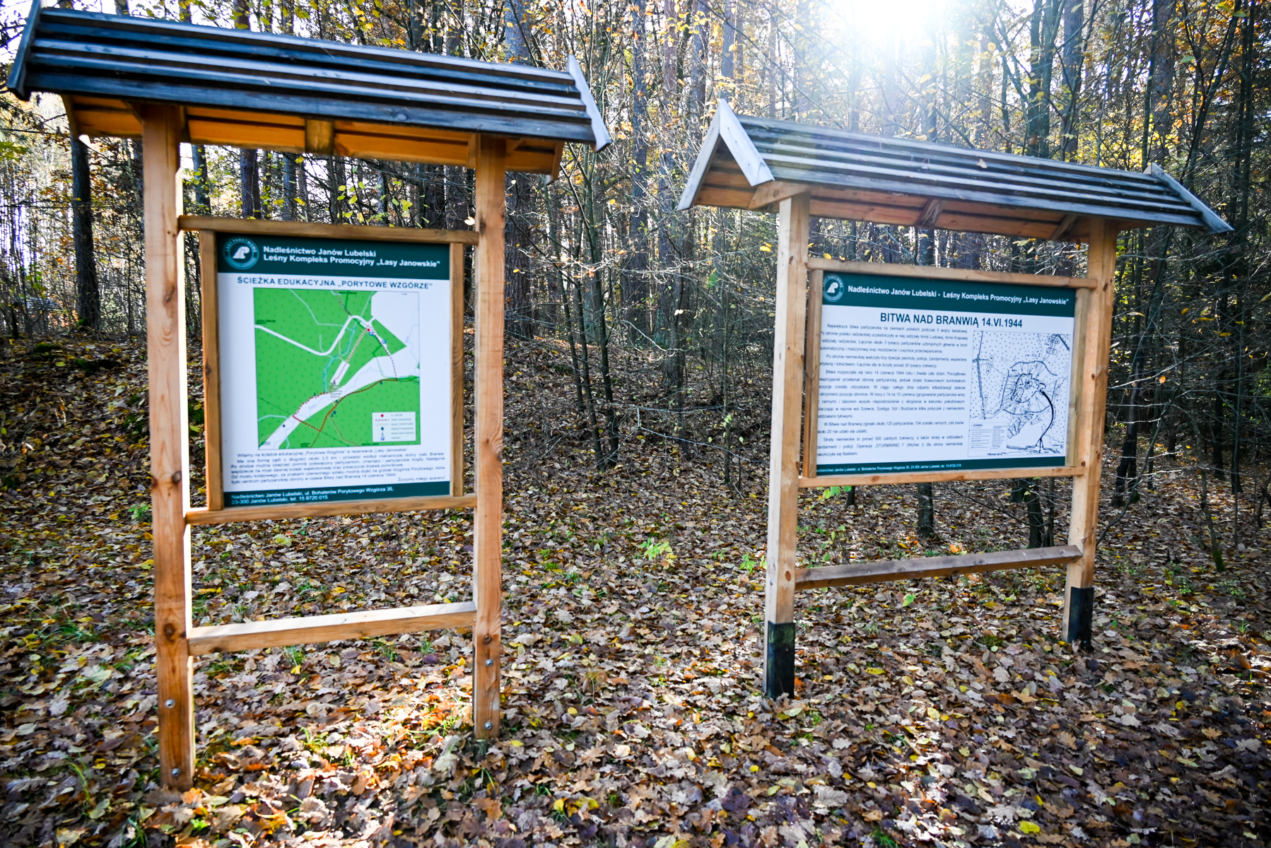 Dwa drewniane tablice informacyjne z daszkami stojące na lesistej, jesiennej ściółce z opadłych liści. W tle drzewa bez liści, światło słoneczne.