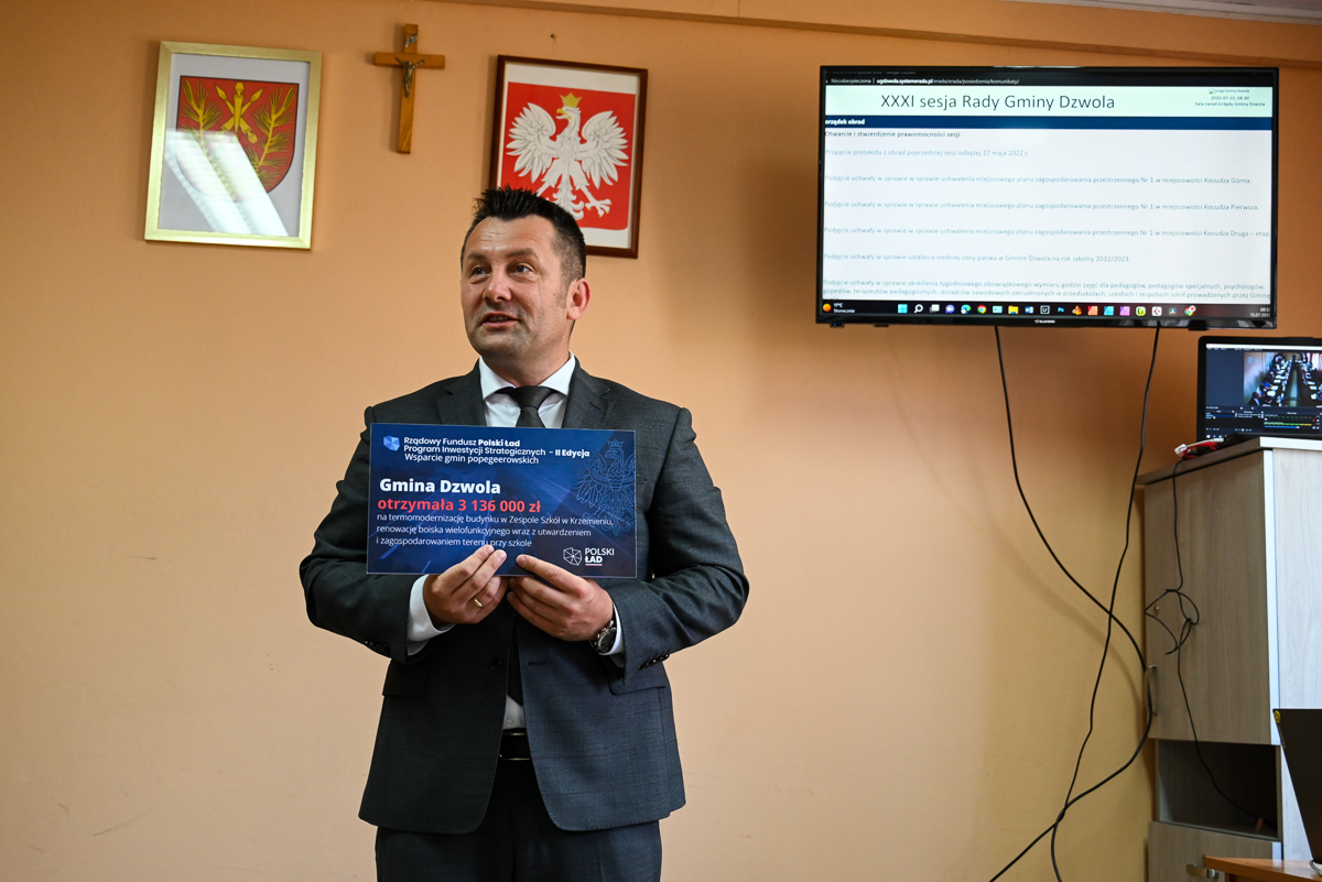 Mężczyzna w ciemnym garniturze trzyma tablicę z napisem o dofinansowaniu dla Gminy Dzwola, za nim pomarańczowa ściana z krzyżem i dwoma herbami, ekran z tekstem sesji rady.