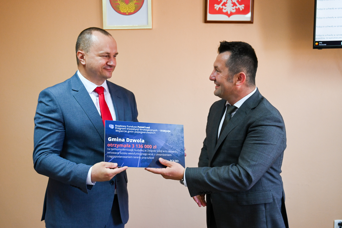 Dwaj mężczyźni w garniturach, jeden z czerwoną krawatem, trzymają niebieską tablicę z tekstem, stoją na tle jasnobrązowej ściany z dwoma herbami i fragmentem monitora.