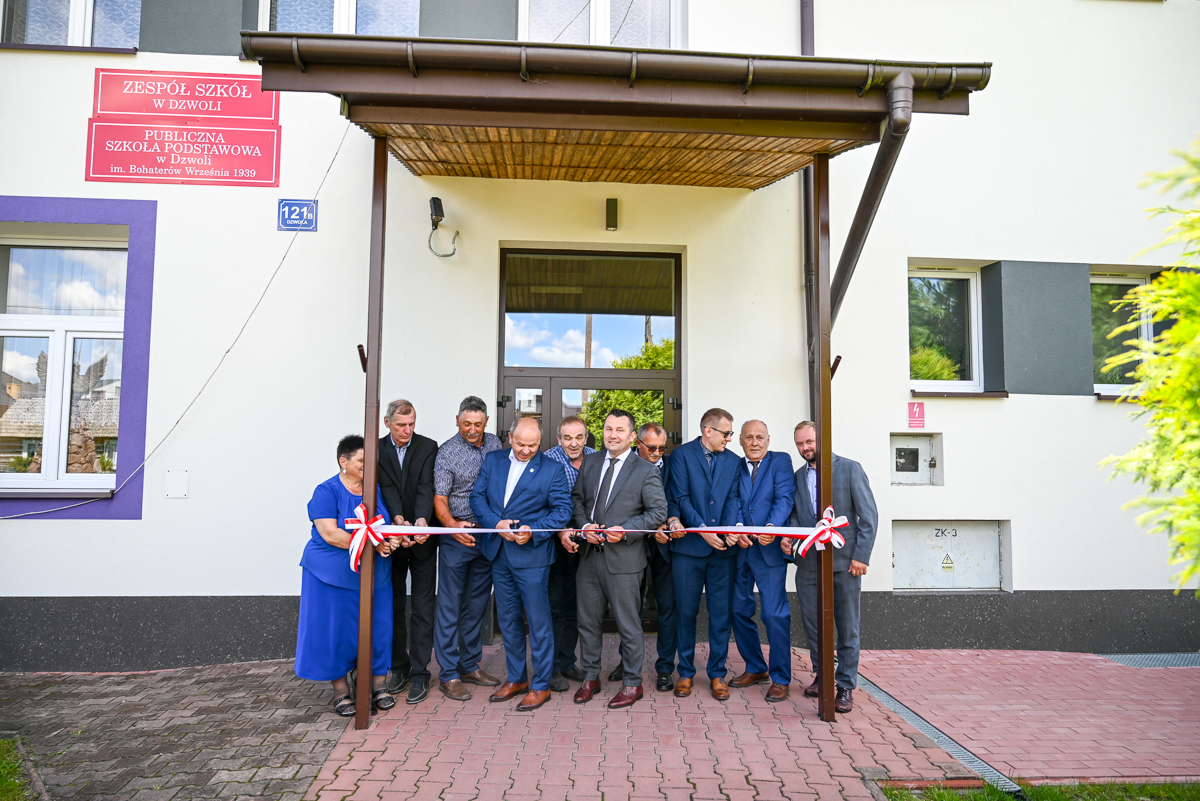 Grupa ludzi w formalnych strojach przecina biało-czerwoną wstęgę przed wejściem do budynku z białą elewacją i fioletowymi ramami okien.