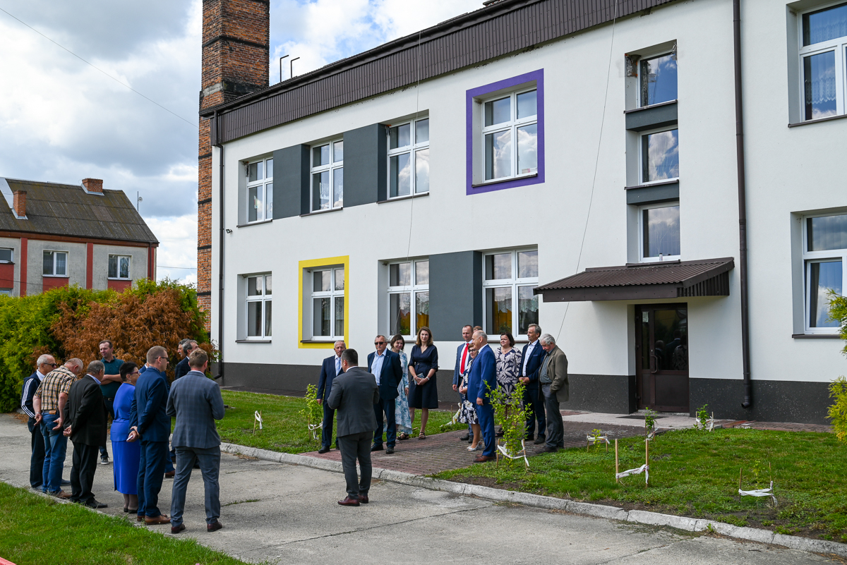 Grupa ludzi stoi na chodniku przed białym budynkiem z kolorowymi ramami okien, trawa i krzewy obok, w tle inny dom i niebo z chmurami.