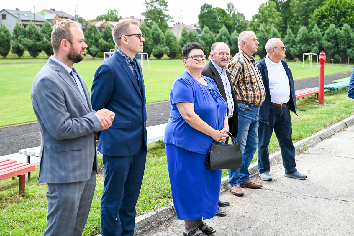 Sześć osób w różnym wieku stoi na zewnątrz na tle zielonej trawy i drzew, pięć mężczyzn i jedna kobieta w niebieskim stroju, kobieta trzyma czarną torebkę.