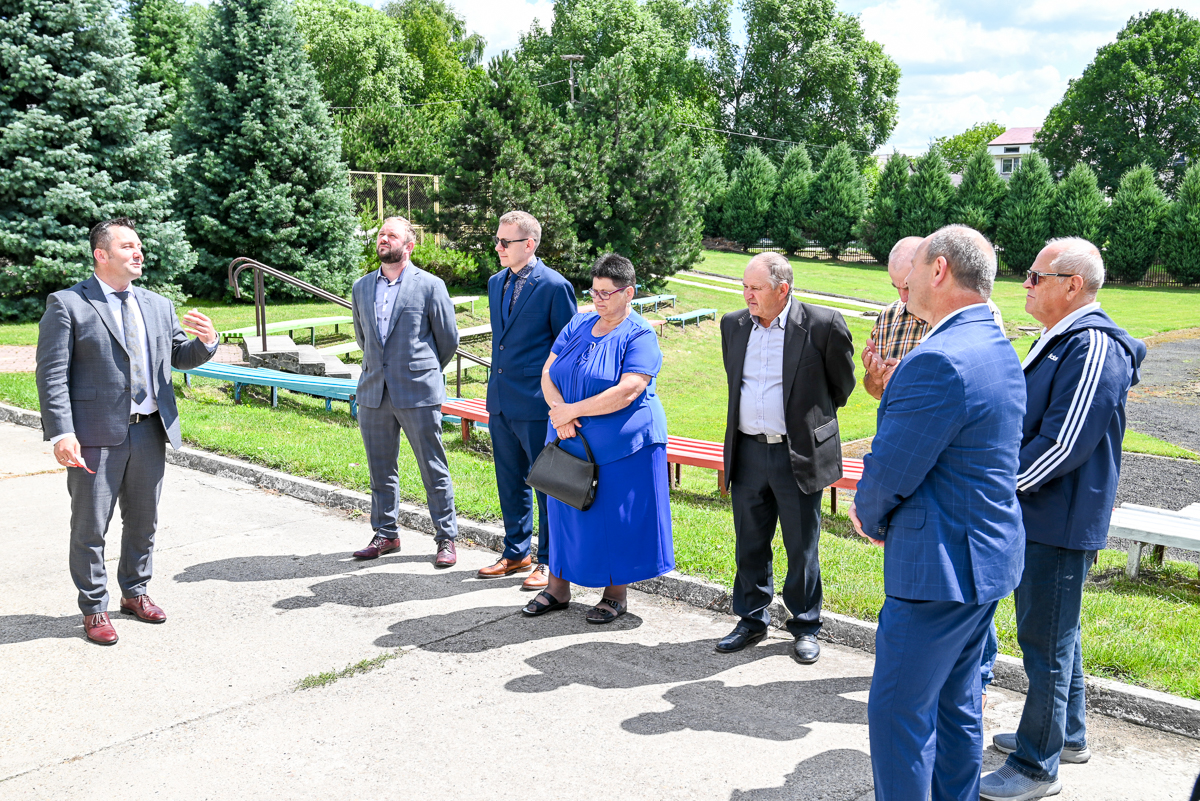 Grupa ośmiu osób na zewnątrz, siedem stoi w rzędzie, jedna gestykuluje, ubrani w garnitury i sukienkę, tło z zielonymi drzewami i trawnikiem.