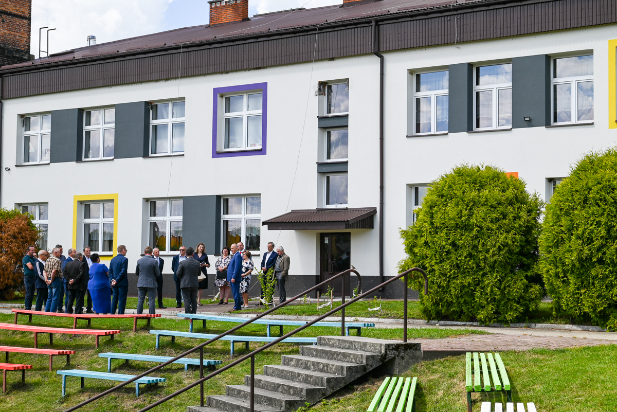 Grupa ludzi na trawniku przed białym budynkiem z kolorowymi ramami okien, obok schody z metalową poręczą, ławki w czerwonym, niebieskim i zielonym kolorze, krzewy.