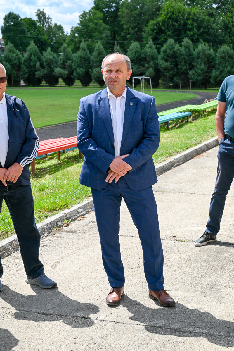 Mężczyzna w niebieskim garniturze i brązowych butach stoi na betonowej ścieżce na tle zielonego boiska i drzew, po bokach fragmenty dwóch innych osób.