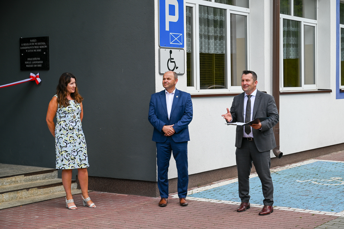 Trzy osoby stojące na chodniku obok budynku z białą ścianą i oknami, dwie osoby w garniturach, kobieta w jasnej sukience, tablica pamiątkowa i znak parkingu dla niepełnosprawnych.