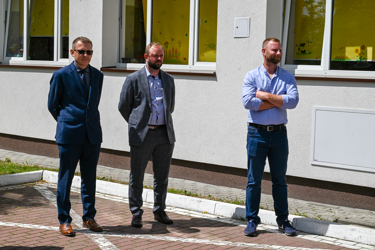 Trzech mężczyzn stoi na chodniku przed jasnym budynkiem z trzema oknami z żółtymi zasłonami. Dwaj po lewej w garniturach, trzeci po prawej w niebieskiej koszuli i ciemnych spodniach.