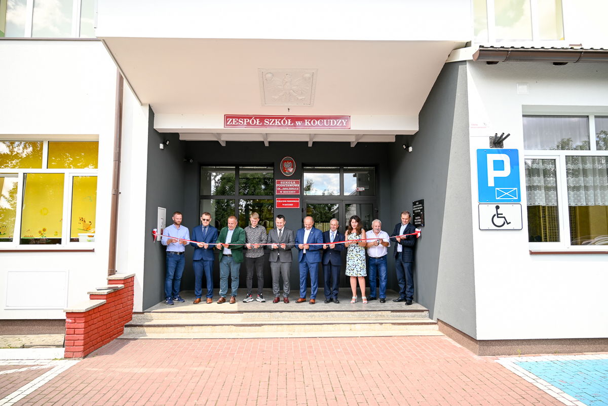 Grupa dziesięciu osób stoi na schodach wejścia budynku, trzymając czerwoną wstęgę, nad wejściem tablica z napisem "ZESPÓŁ SZKÓŁ w KOCUDZY" i symbol orła.