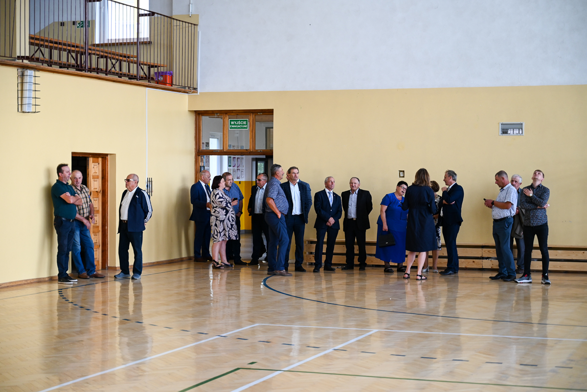 Grupa ludzi w różnym wieku w jasnej sali gimnastycznej z drewnianą podłogą i żółto-białymi ścianami, niektórzy rozmawiają, inni stoją osobno.