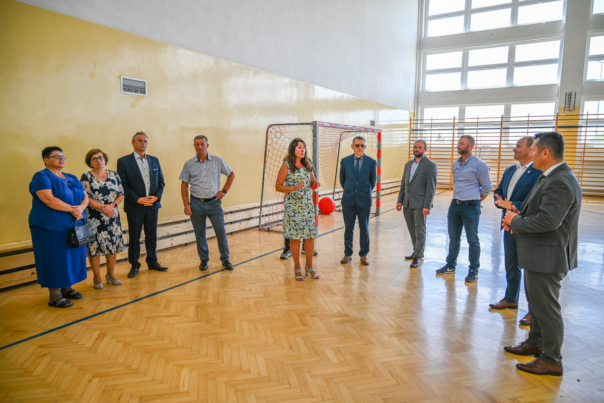 Grupa jedenastu osób stojących w dużej jasnej sali gimnastycznej z drewnianą podłogą i siatką do piłki ręcznej, w większości ubranych w garnitury i sukienki.