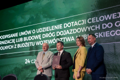 Cztery osoby w garniturach stoją na tle zielonego ekranu z tekstem w języku polskim, jedna trzyma dokument.