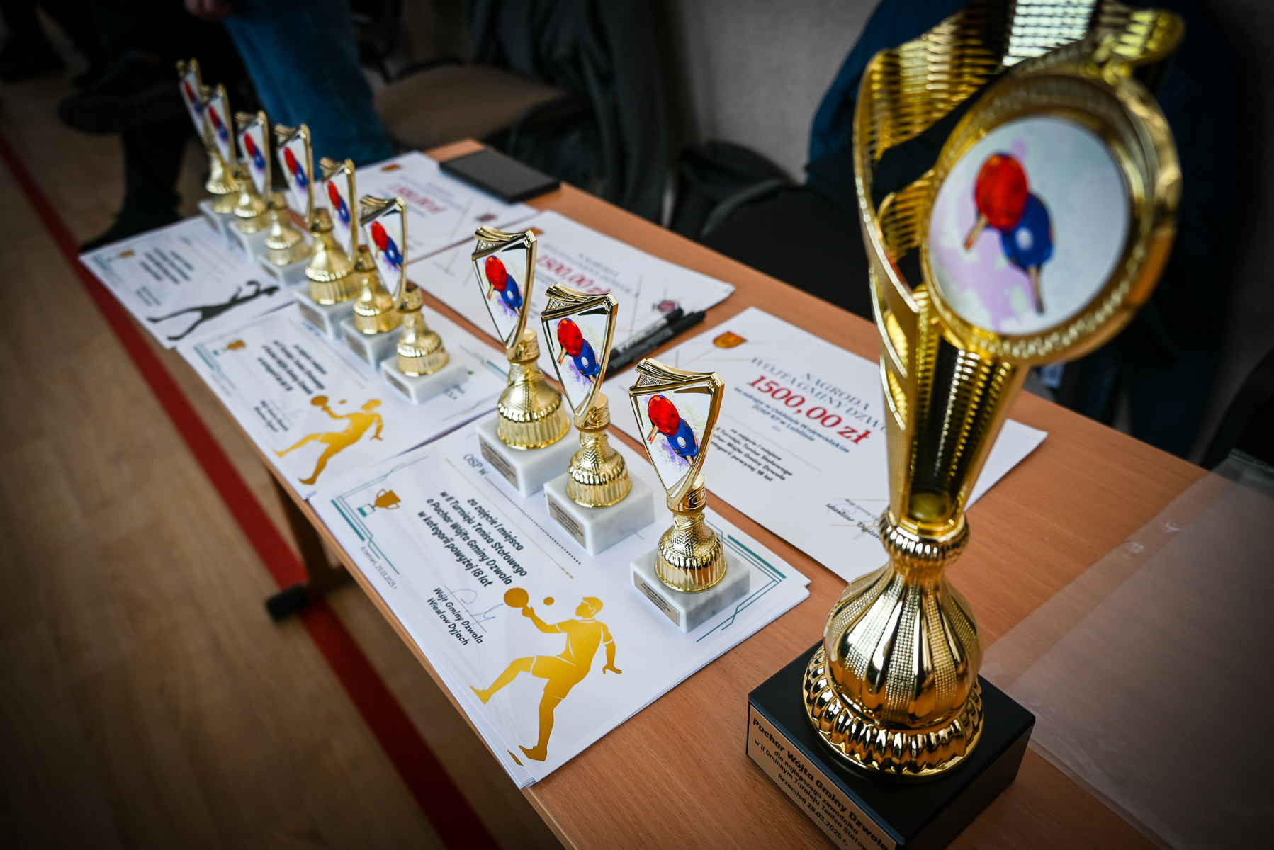 Złote puchary i statuetki do tenisa stołowego na drewnianym stole, pod nimi białe dyplomy z żółtymi sylwetkami grających ludzi.