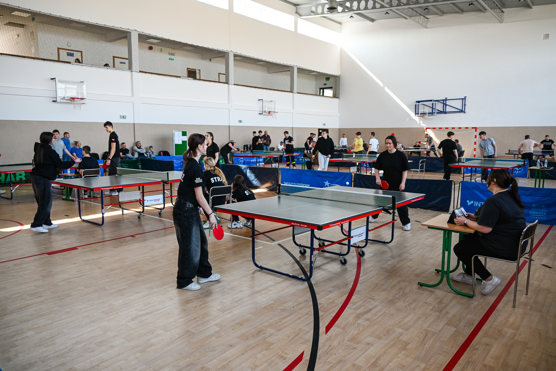 Ludzie grający i obserwujący tenis stołowy w jasnej sali gimnastycznej z drewnianą podłogą, stoły do ping-ponga, czerwone rakietki, niebieskie i czarne bariery.