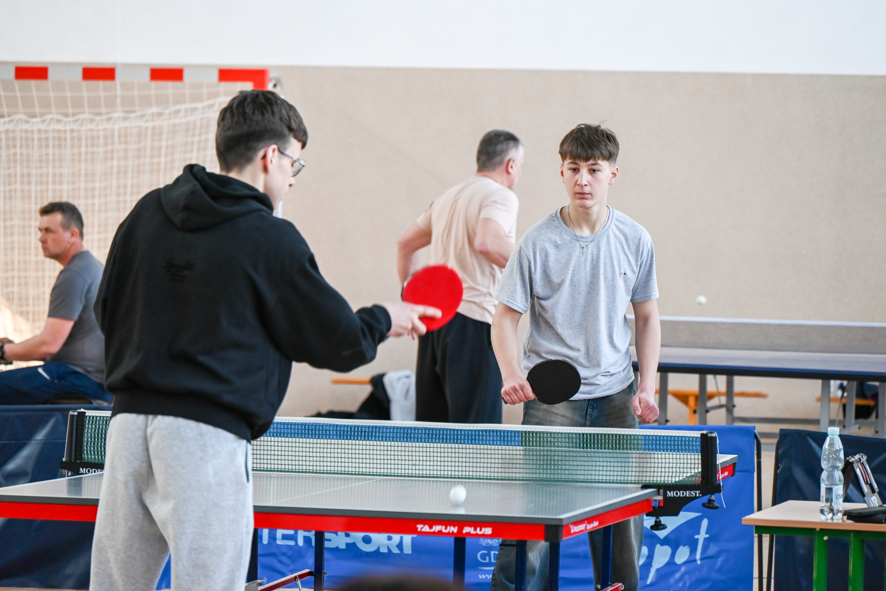 Dwóch młodych mężczyzn gra w tenisa stołowego przy zielonym stole, jeden w czarnej bluzie i jasnych spodniach, drugi w szarej koszulce i jeansach, w tle siatka bramki.