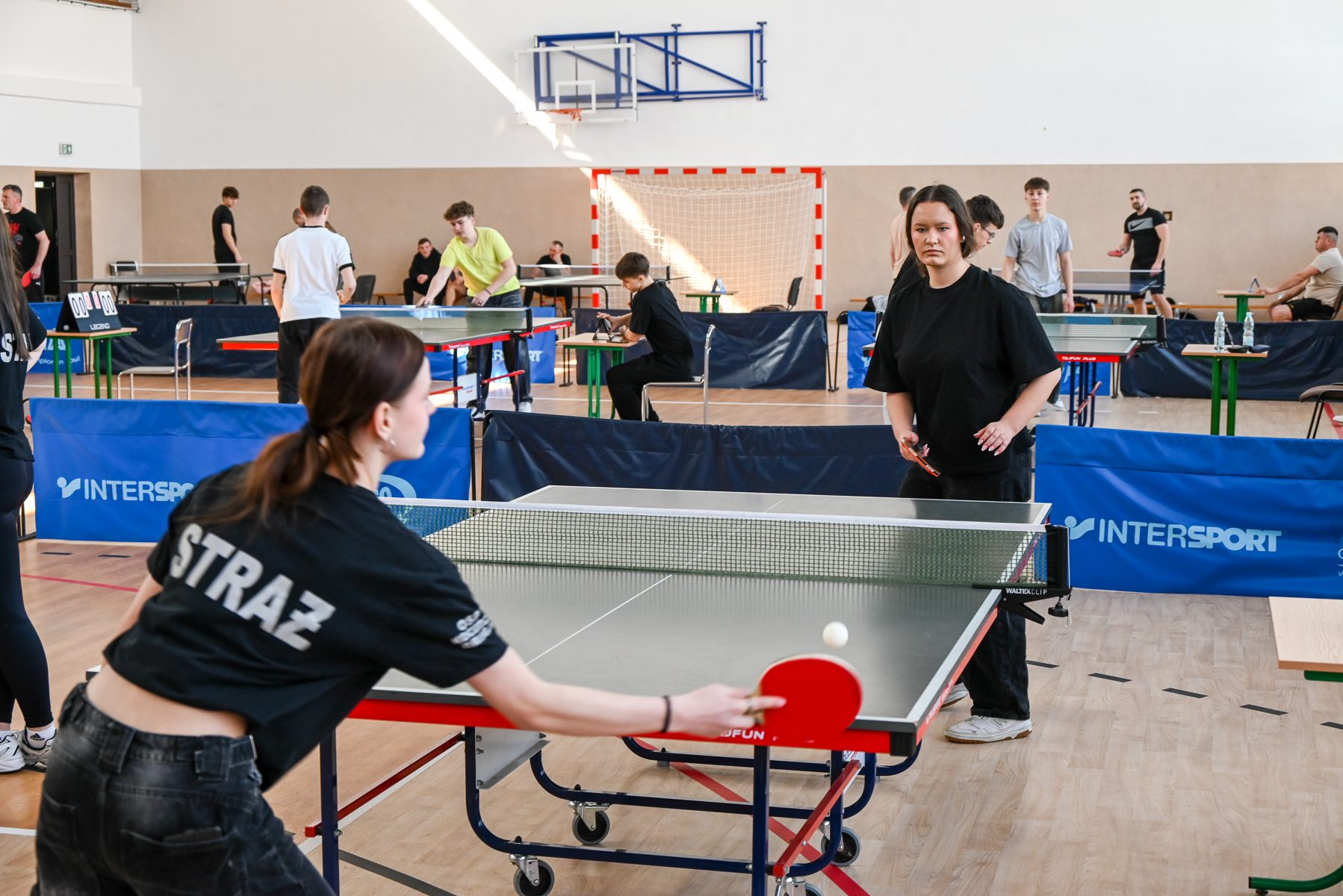 Hala sportowa z kilkoma stołami do tenisa stołowego, osoby grające w piłkę, niebieskie bandy z napisem INTERSPORT, drewniana podłoga.