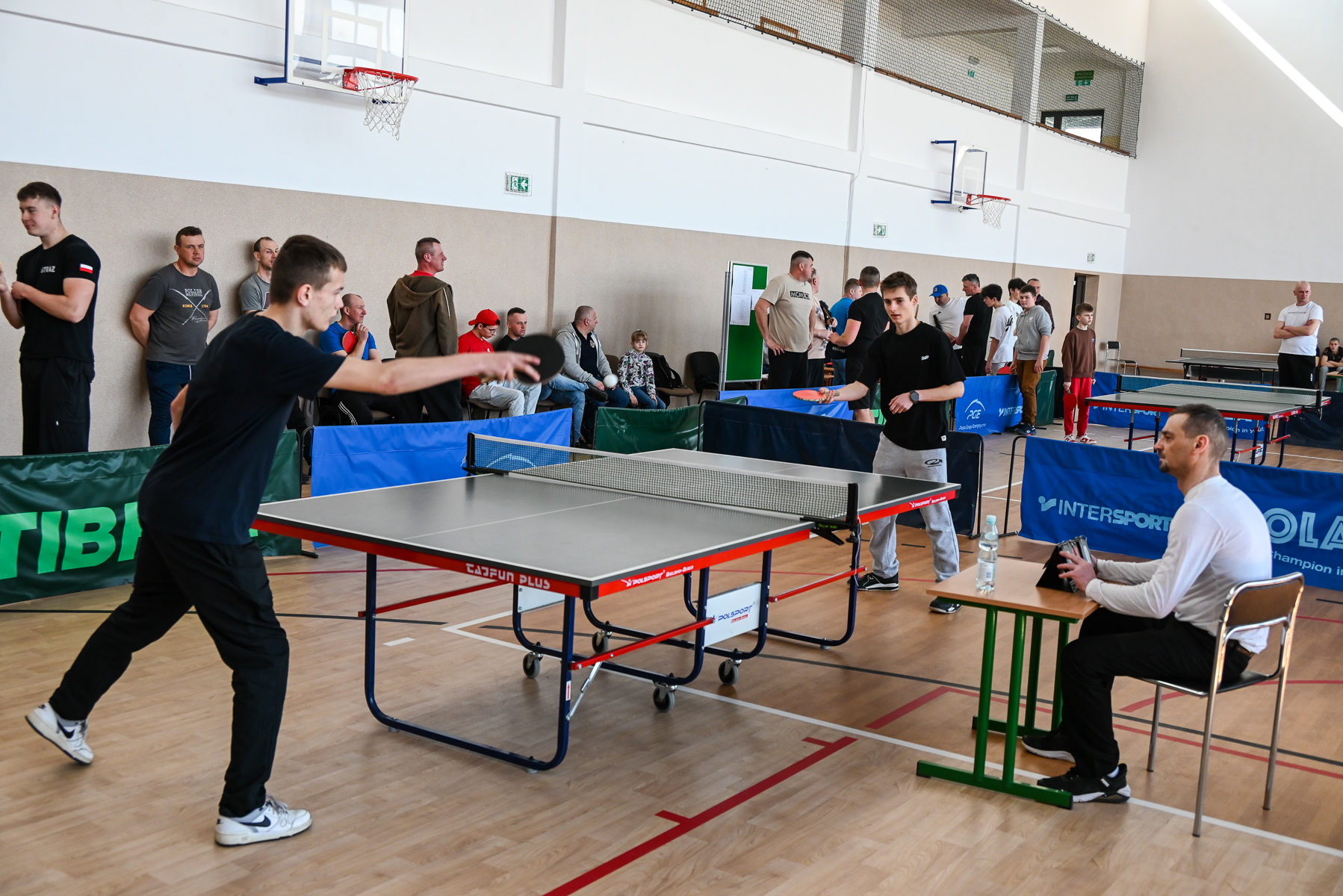 Dwóch młodych mężczyzn gra w tenisa stołowego przy szarej składanej planszy, sędzia siedzi przy stoliku obok, w tle widoczna grupa osób obserwujących rozgrywkę w hali sportowej.
