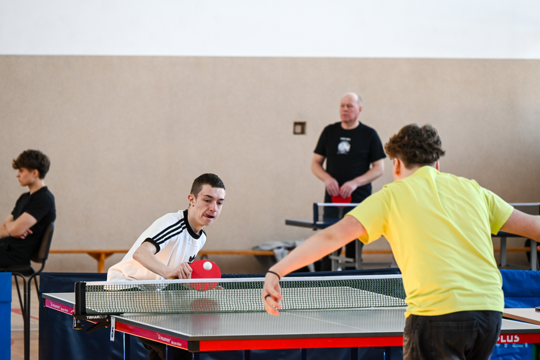 Dwóch chłopców gra w tenisa stołowego, jeden w białej koszulce, drugi w żółtej, w tle dwóch mężczyzn, jeden siedzi, drugi stoi.
