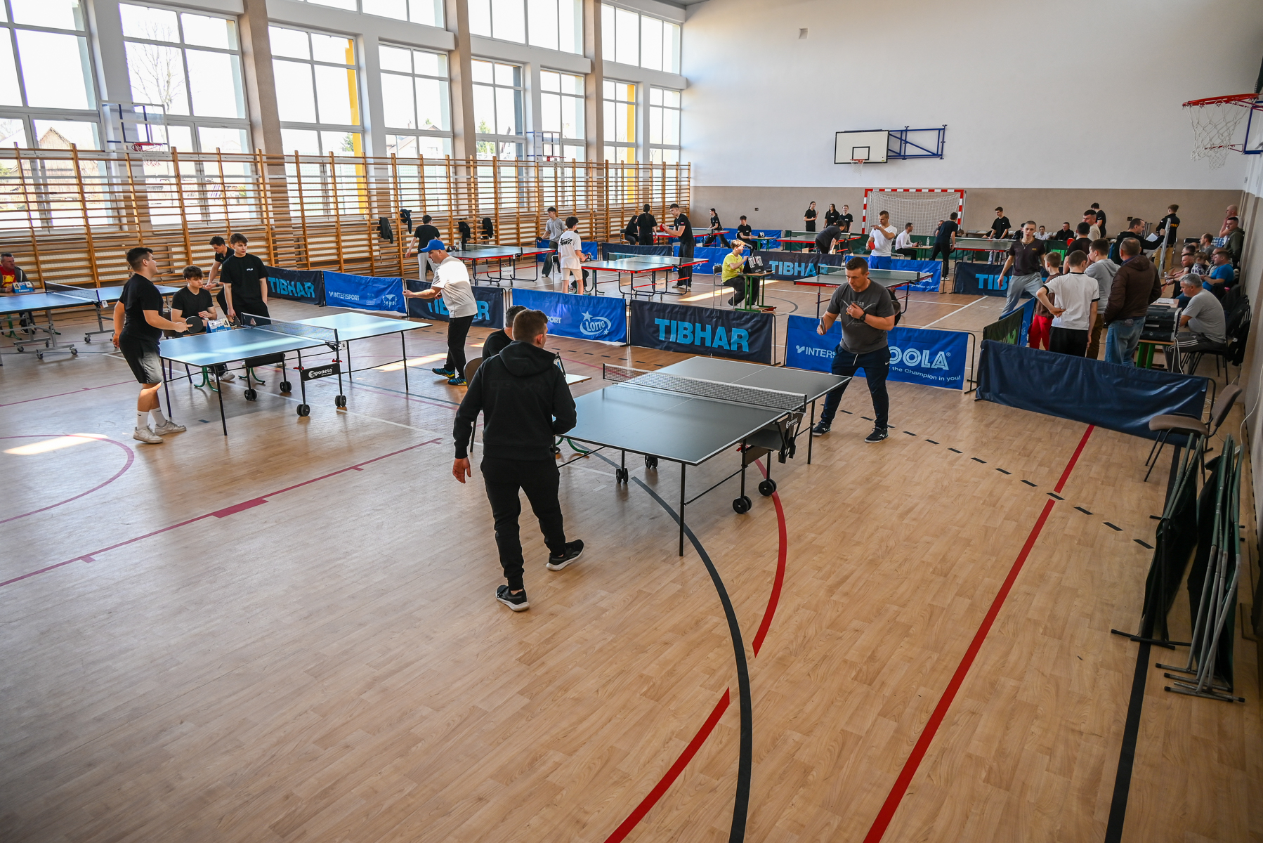 Hala sportowa z jasną drewnianą podłogą, dużymi oknami, kilkoma stołami do tenisa stołowego i grupami grających mężczyzn oraz chłopców.